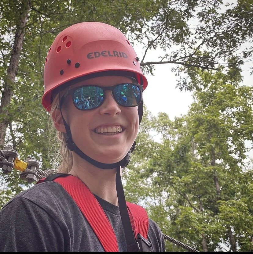 Woman smiling outdoors wearing a red safety helmet, black sunglasses, a black shirt, and red harness set against a background of green trees.