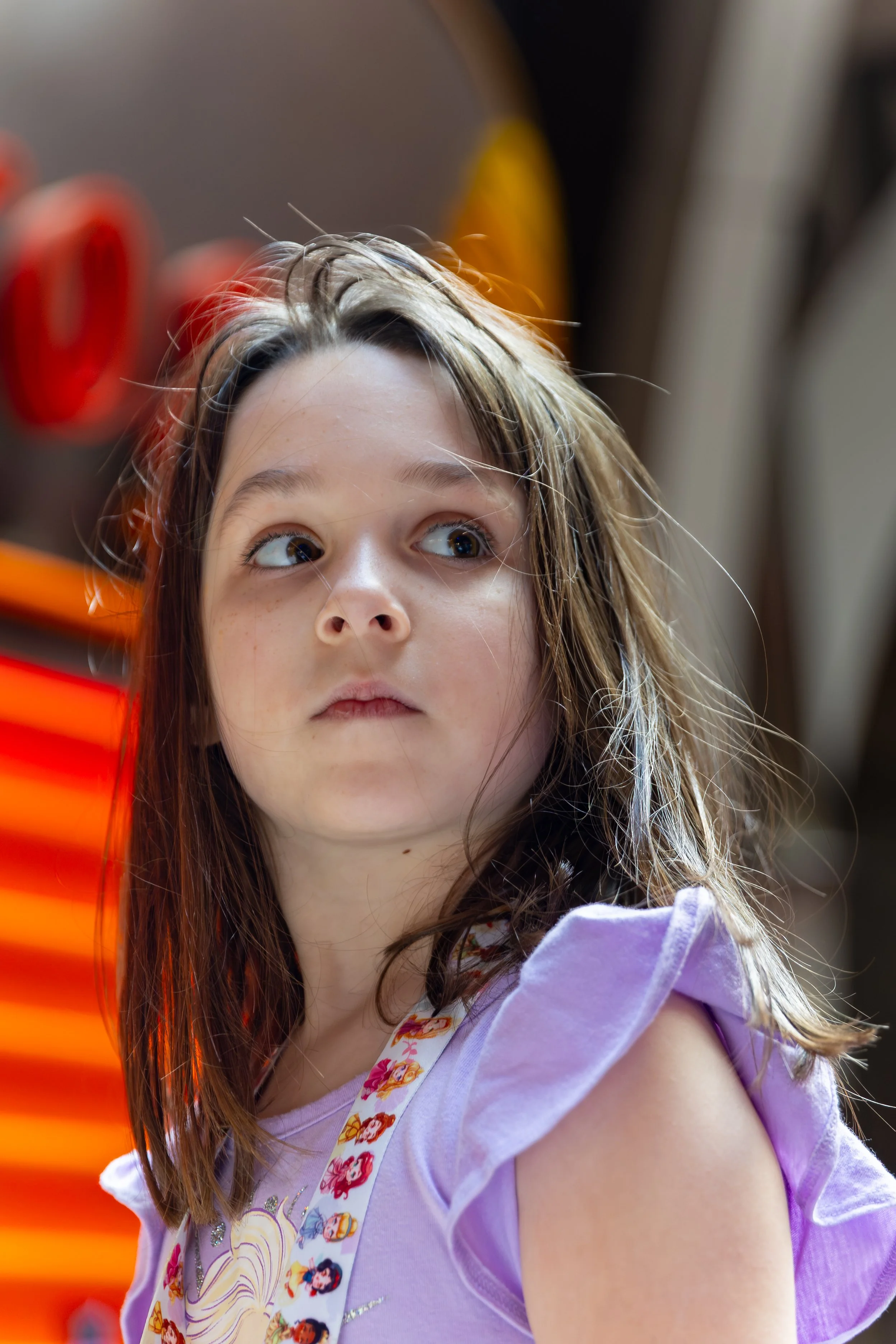 A young girl with long brown hair, wearing a purple sleeve shirt and a Minnie Mouse themed suspenders, sitting in front of a blurred background with orange and black colors.