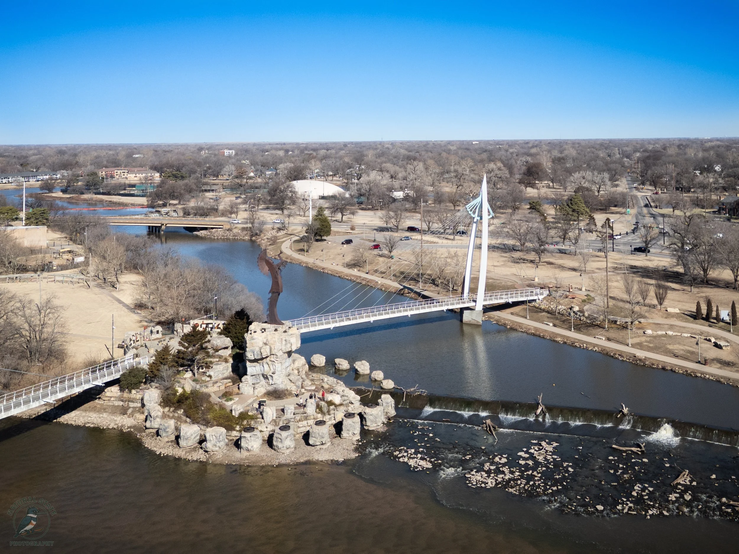 View of a cityscape with a river running through a park, a white bridge, and a modern, sail-shaped architectural structure under a clear blue sky.