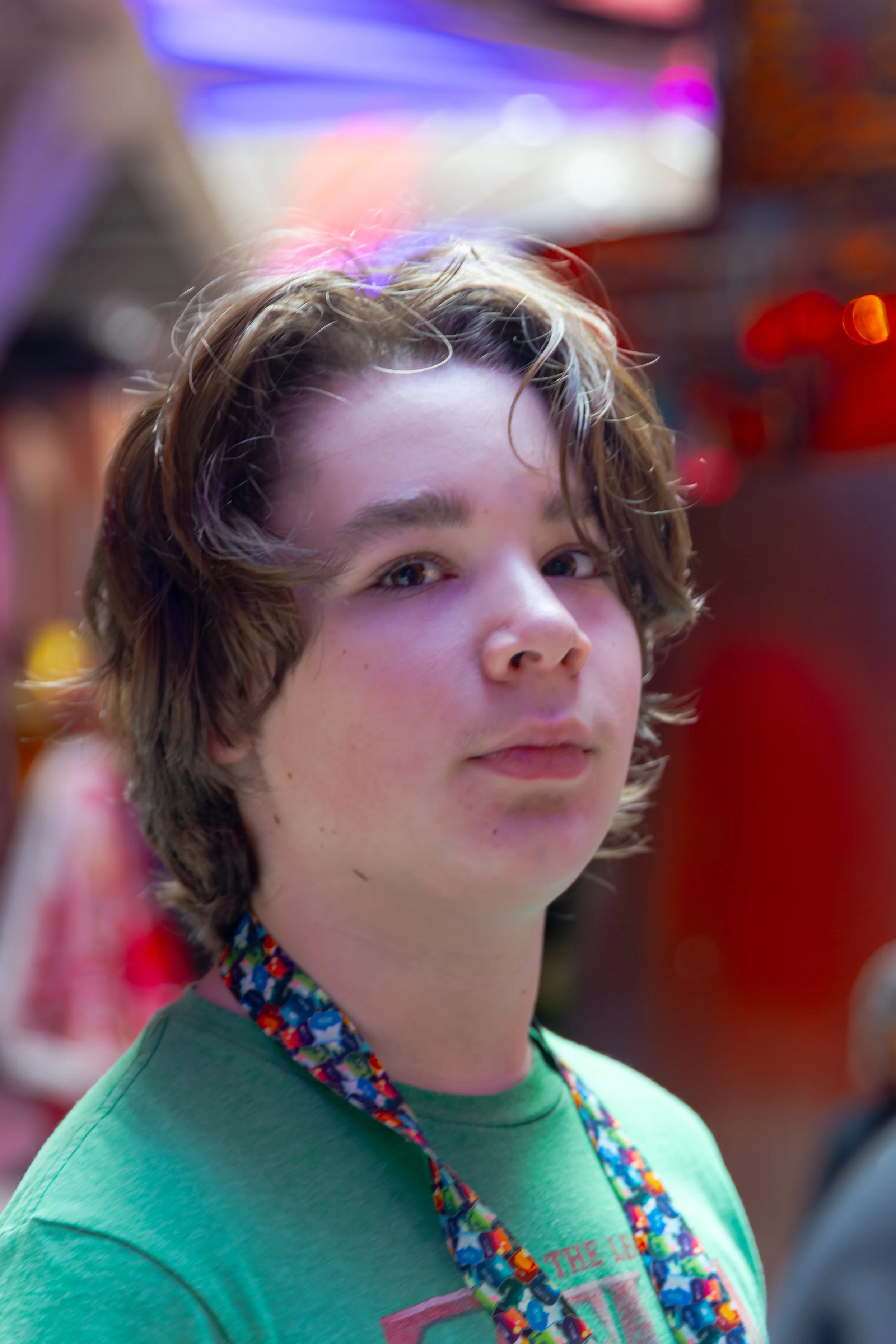 A young person with short, wavy hair, wearing a green T-shirt and a colorful lanyard, smiling slightly with a blurred background of vibrant lights.