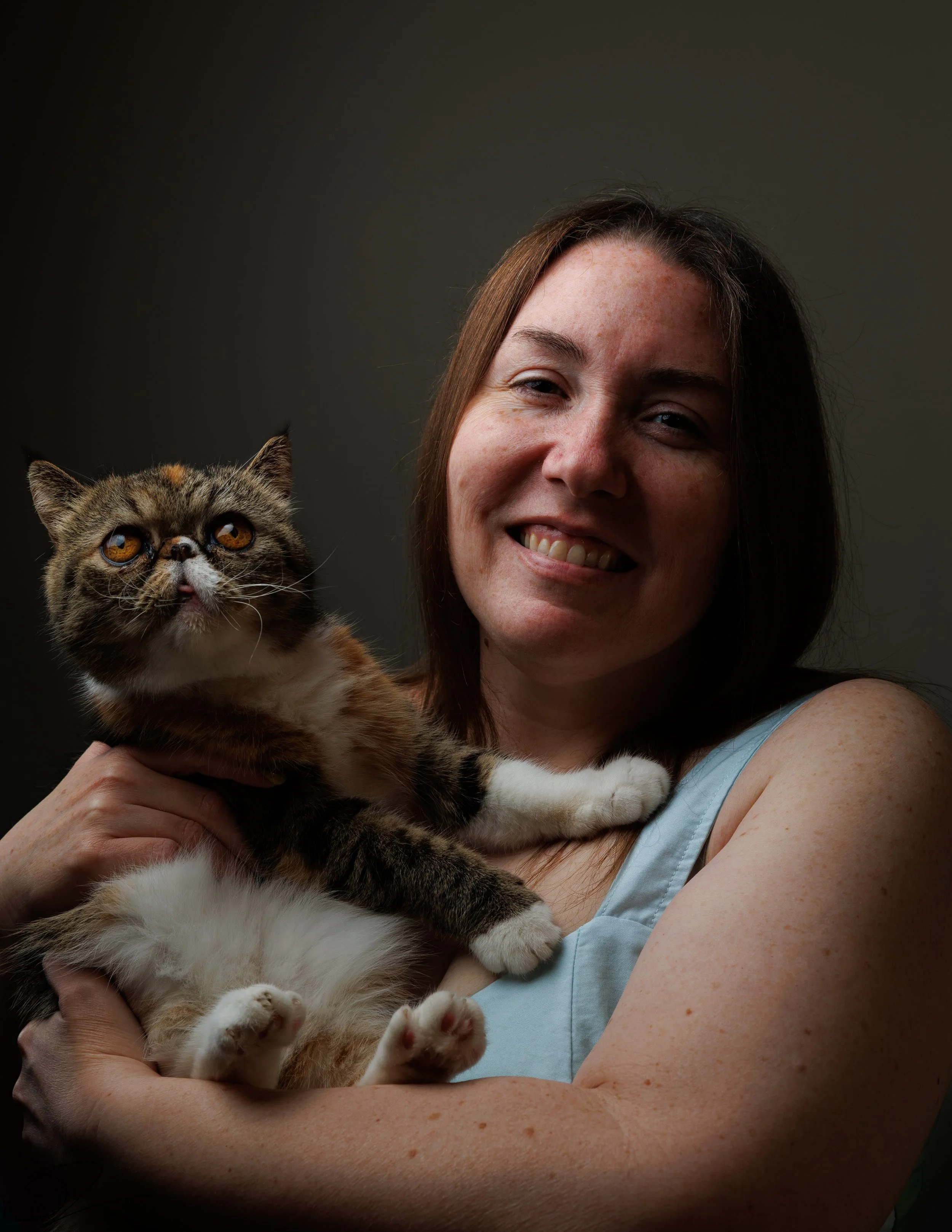 A woman with brown hair and freckles holding a calico cat with wide eyes, set against a dark background.