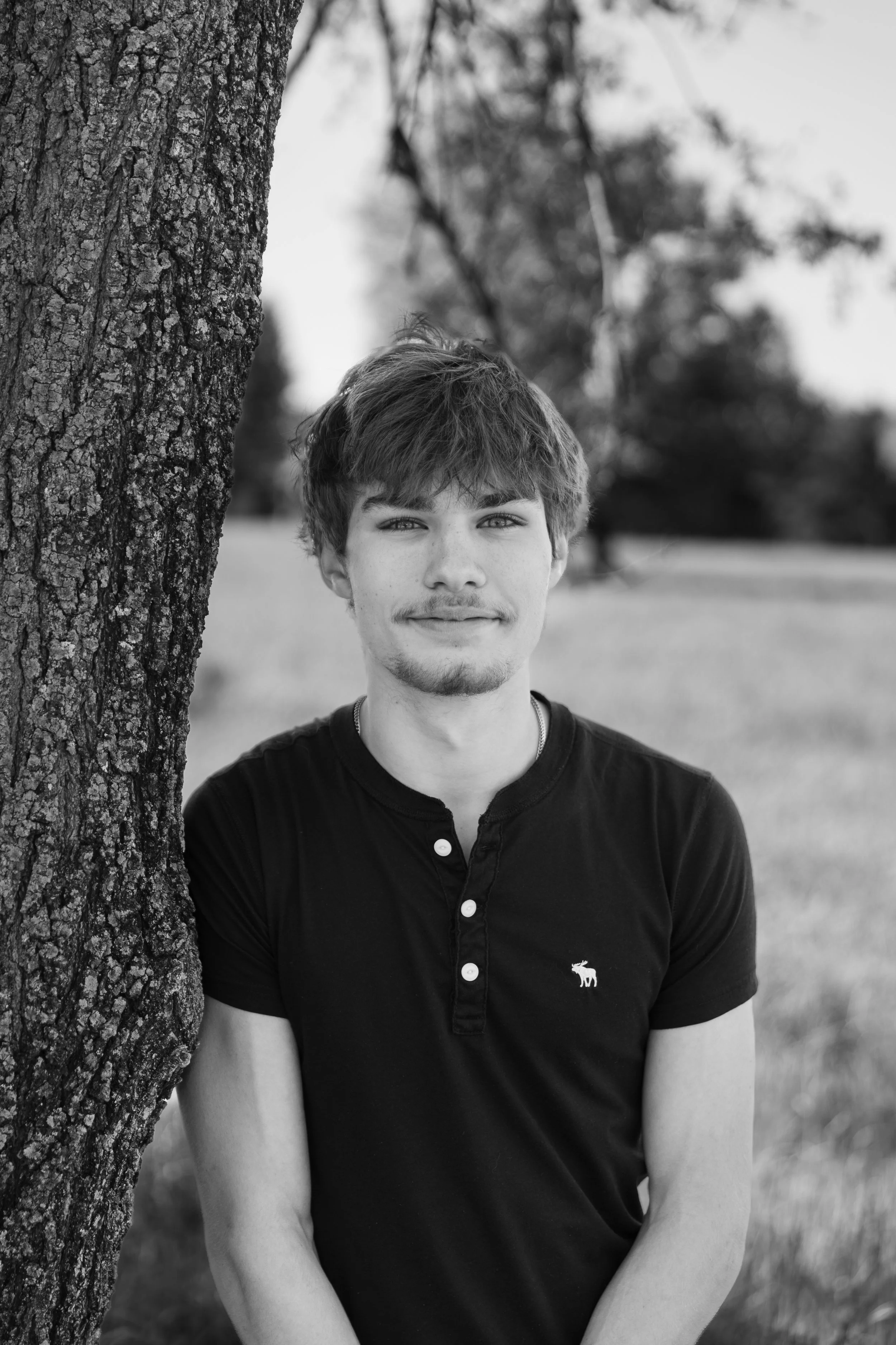 Black and white photo of a young man with light-colored hair, standing outdoors next to a tree, with a blurred natural background.