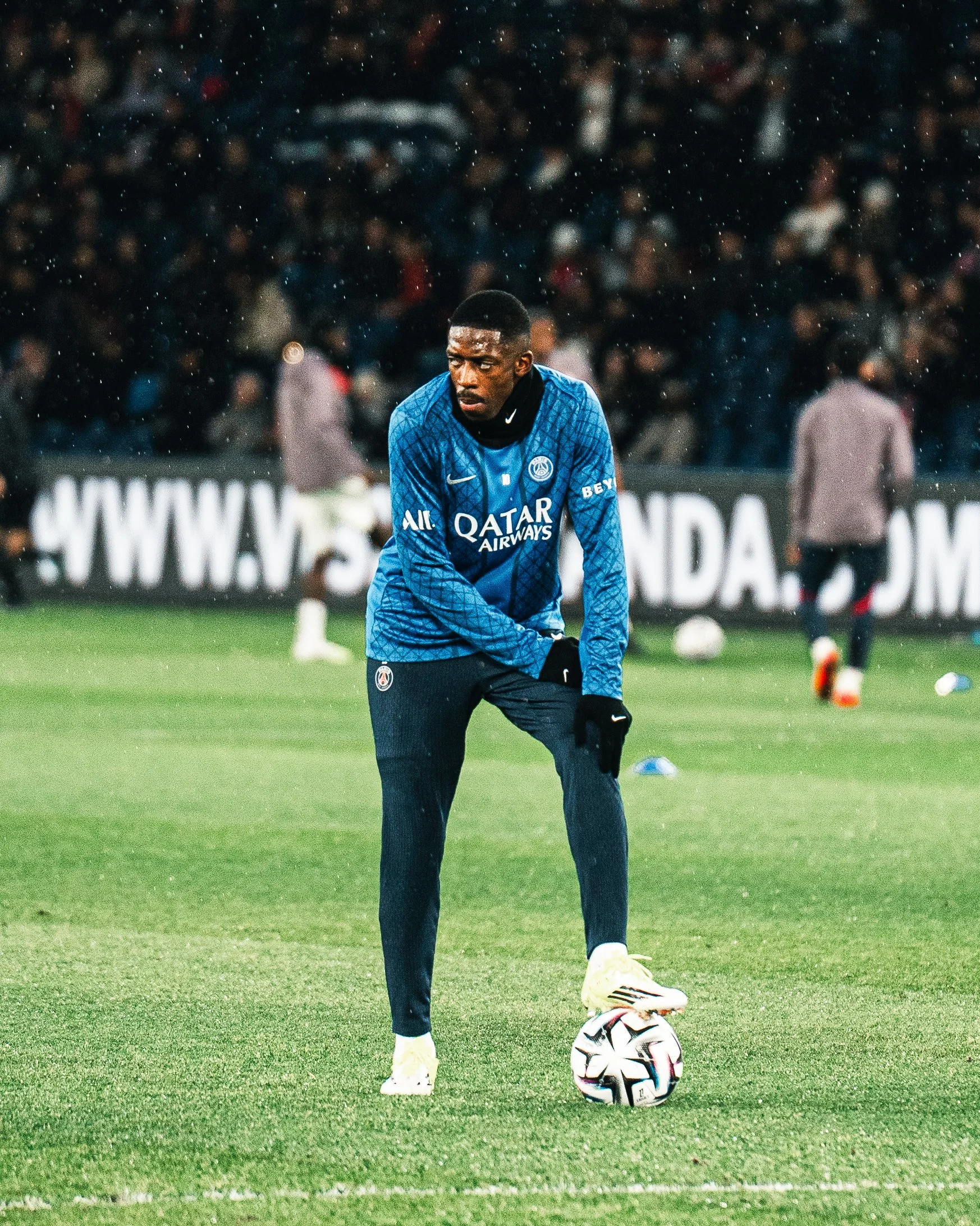 Un joueur de football en entraînement, portant un maillot bleu de Paris Saint-Germain, se prépare à jongler avec un ballon sur le terrain.