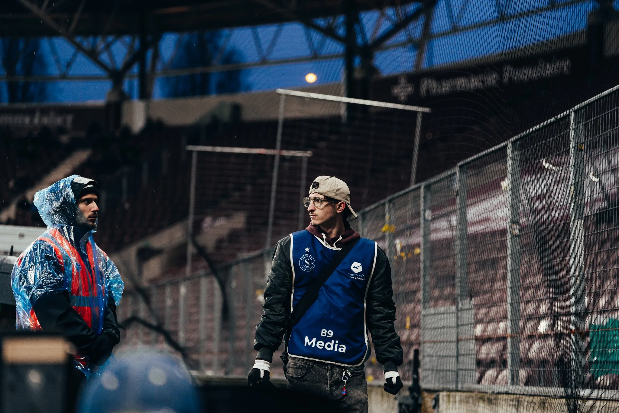 Deux jeunes hommes discutent près d'une cage de football en plein air, par temps de pluie, avec un ciel sombre et des structures métalliques au-dessus.