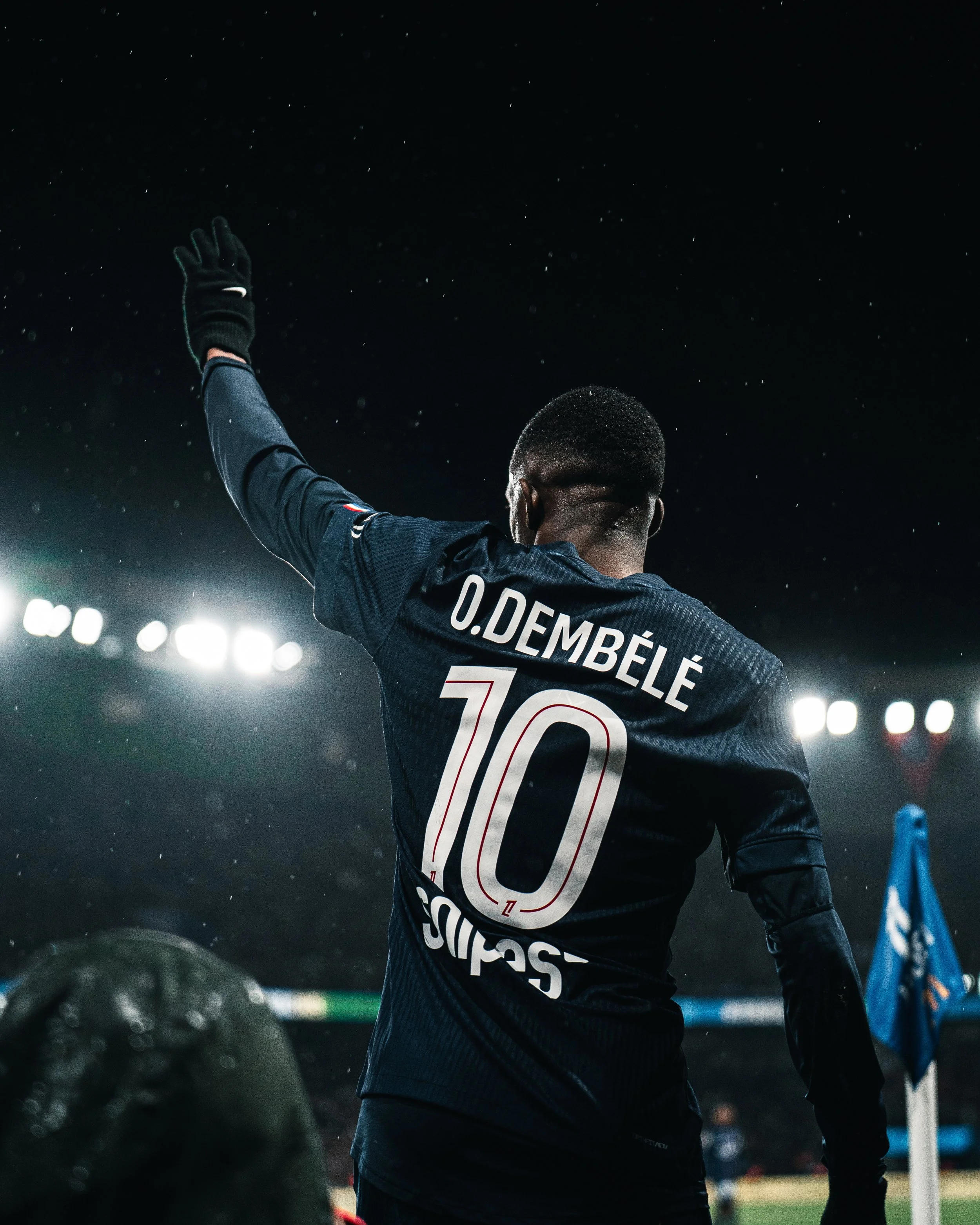 Footballeur de dos, portant le maillot avec le nom D. DEMBÉLÉ et le numéro 10, levant la main dans un stade sous la pluie la nuit.