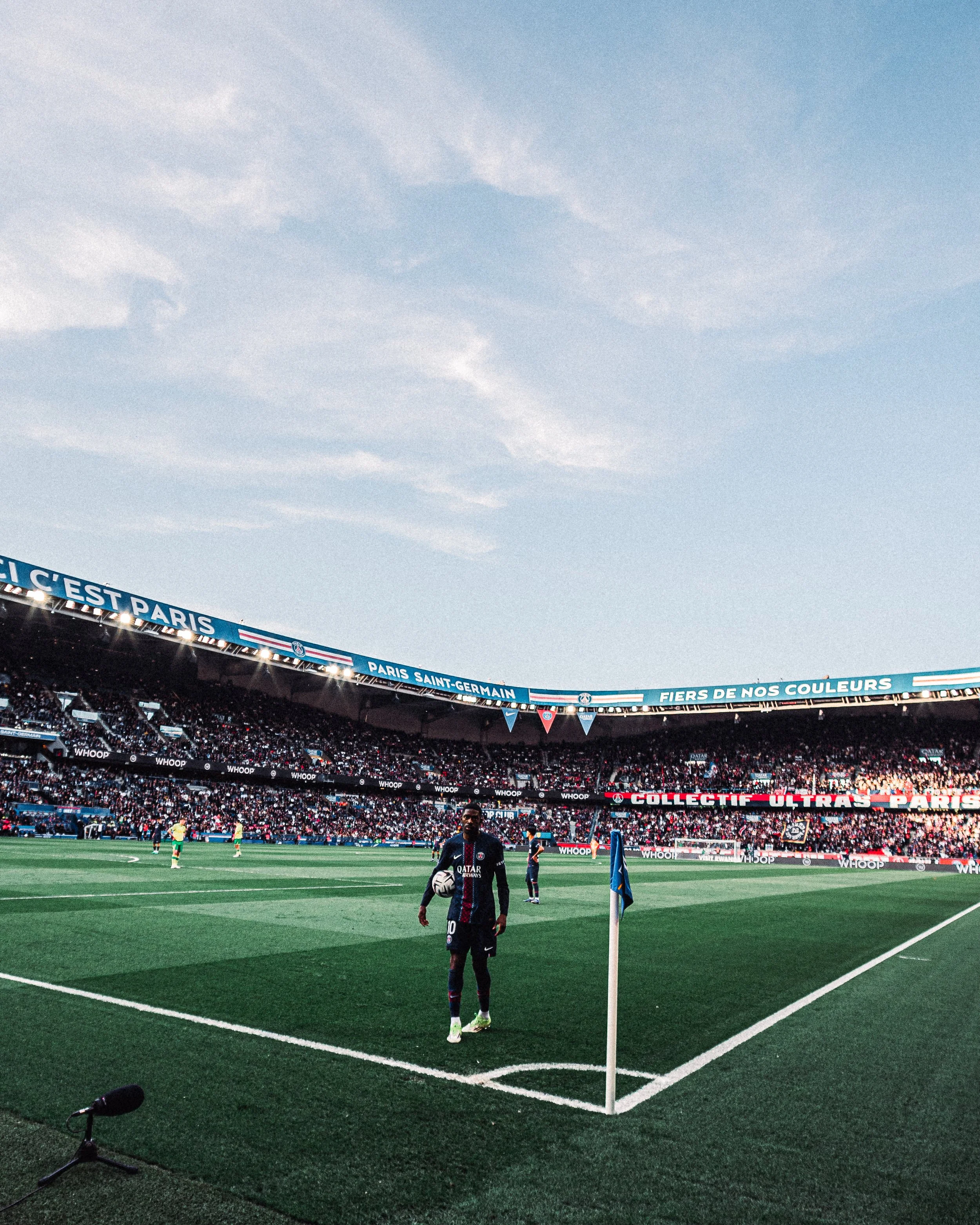 Un joueur de football portant un maillot du Paris Saint-Germain se tient sur le terrain, près du drapeau de corner, lors d'un match avec une foule dans le stade et un ciel partiellement nuageux.
