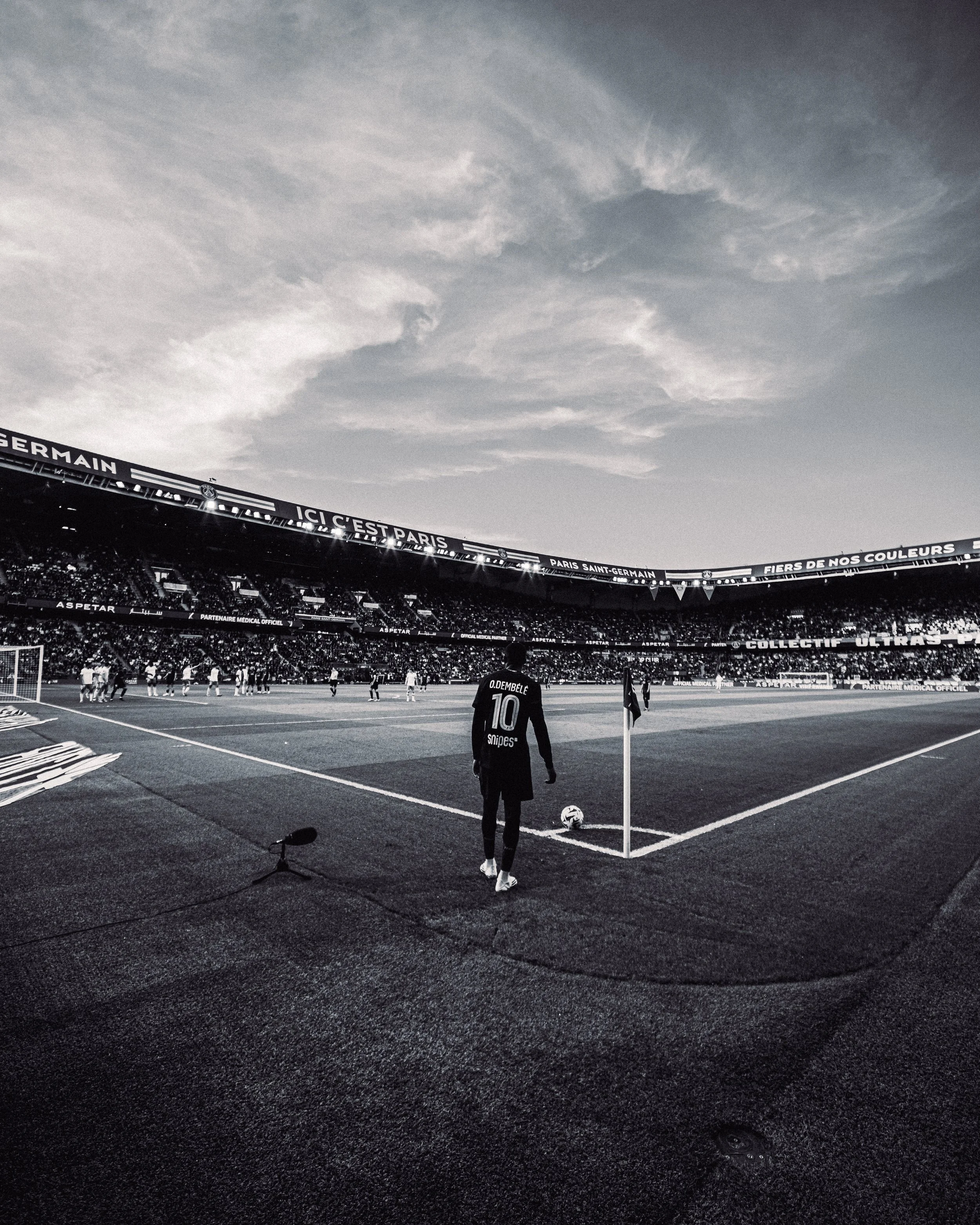 Vue d'un stade de football avec un joueur en arrière-plan portant le numéro 10, la pelouse, des spectateurs, et une partie de la pelouse avec ligne de touche.