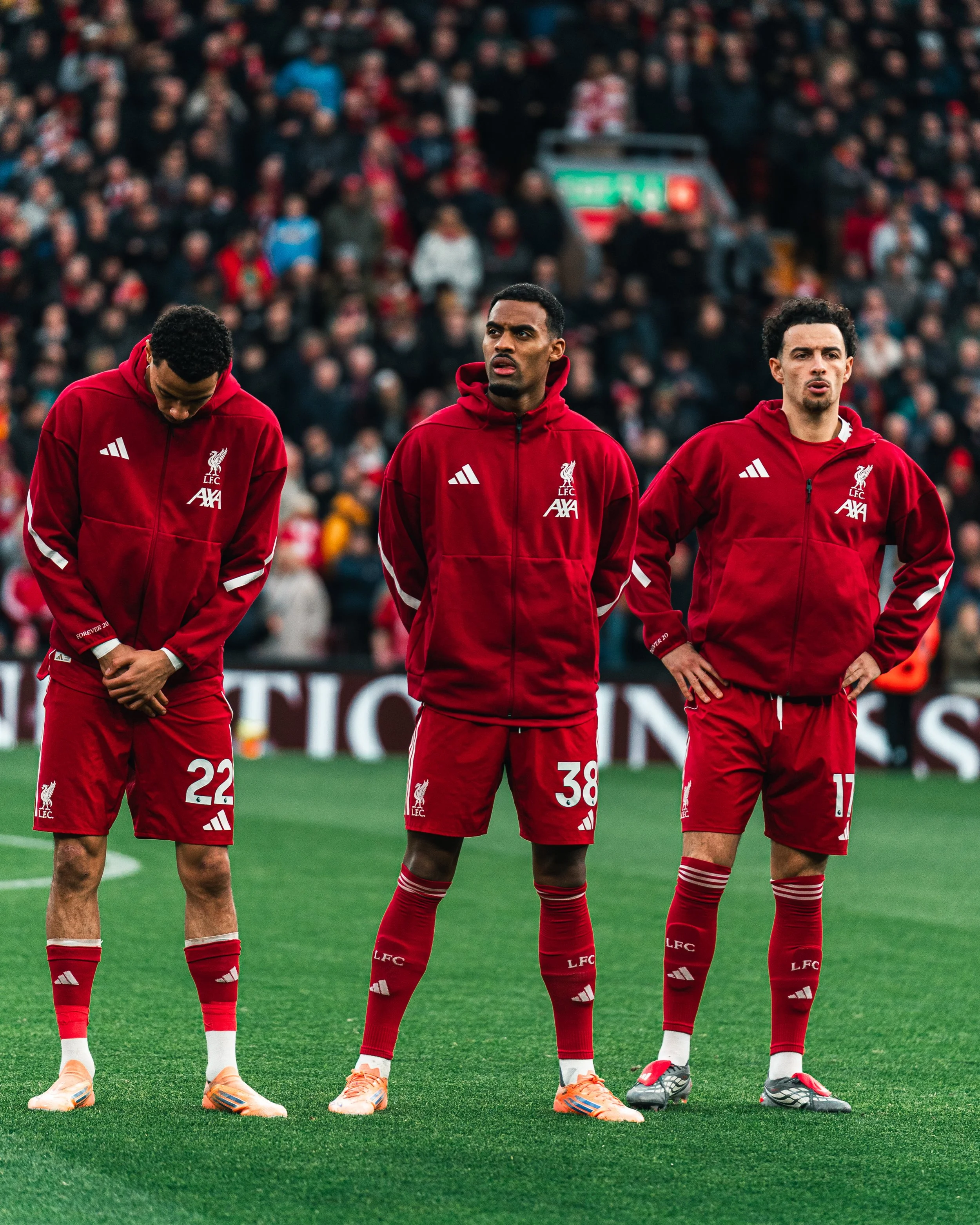 Trois joueurs de football en tenue rouge, portant les logos de Liverpool et d'Axa, se tiennent sur le terrain avant un match, avec un public dans les gradins en arrière-plan.