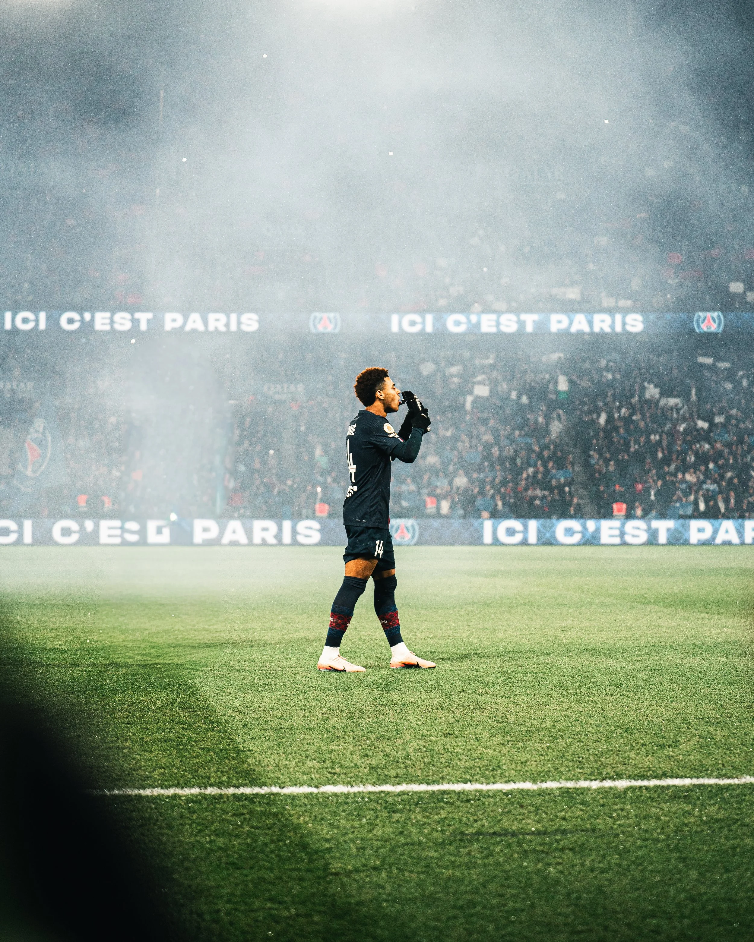 Un joueur de football en uniforme noir est debout sur le terrain, tenant un microphone à la main. En arrière-plan, un panneau indique 'ICI C'EST PARIS' et il y a un grand stade rempli de spectateurs.
