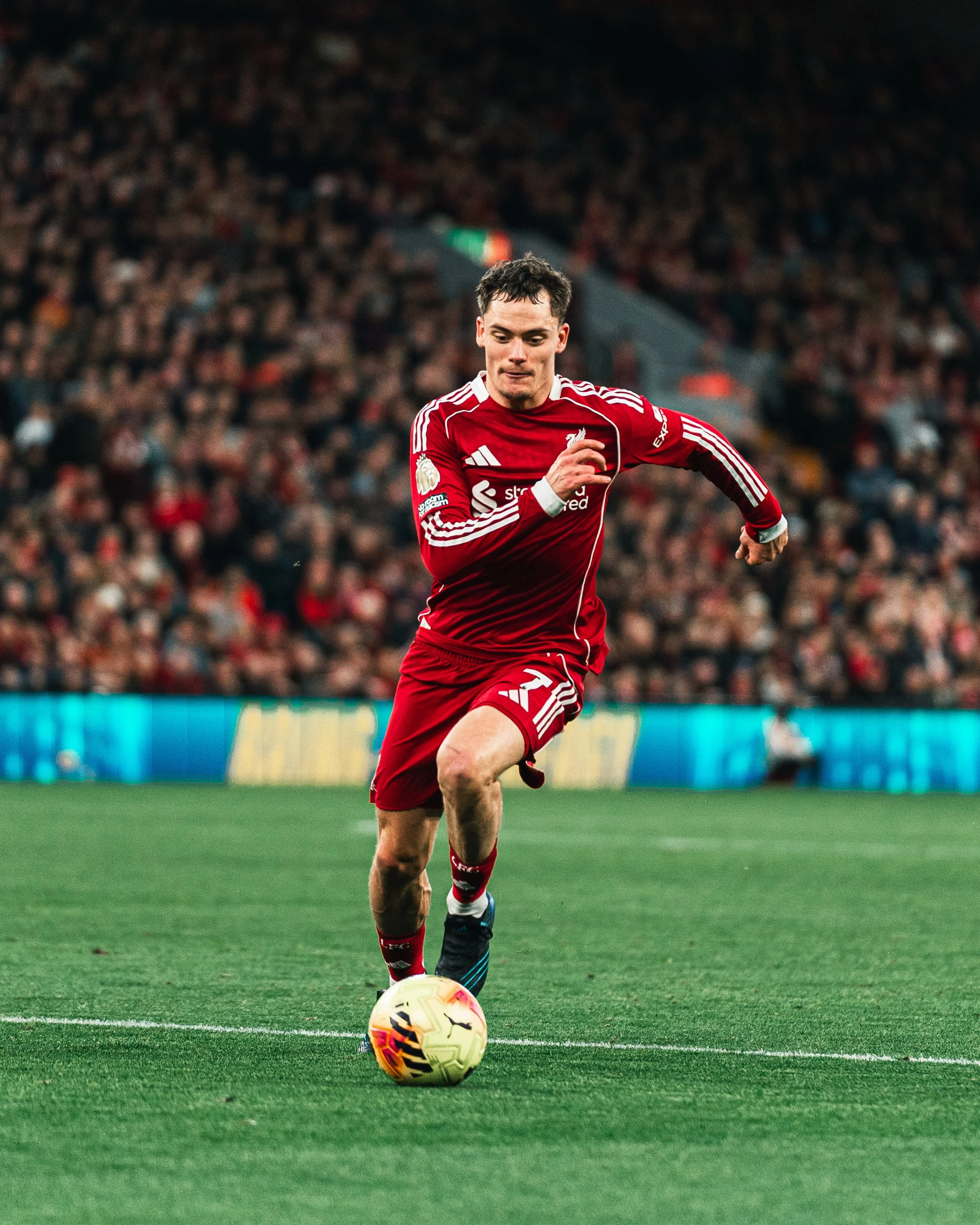 Un joueur de football en maillot rouge en train de courir avec le ballon sur le terrain devant une foule nombreuse.