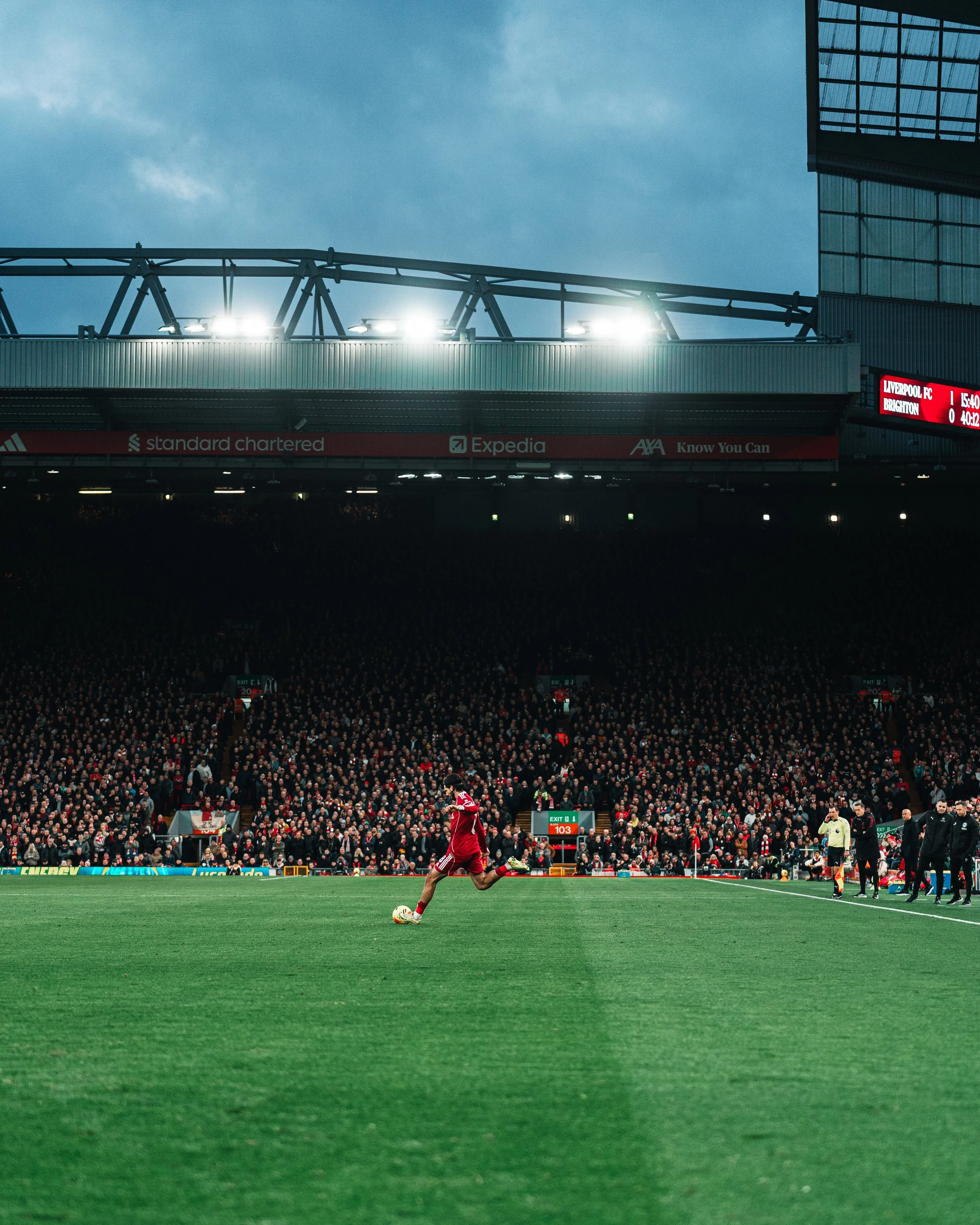 Une scène de match de football dans un grand stade, avec un joueur en tenue rouge frappant le ballon, sous un ciel nuageux, avec une foule nombreuse dans les gradins.