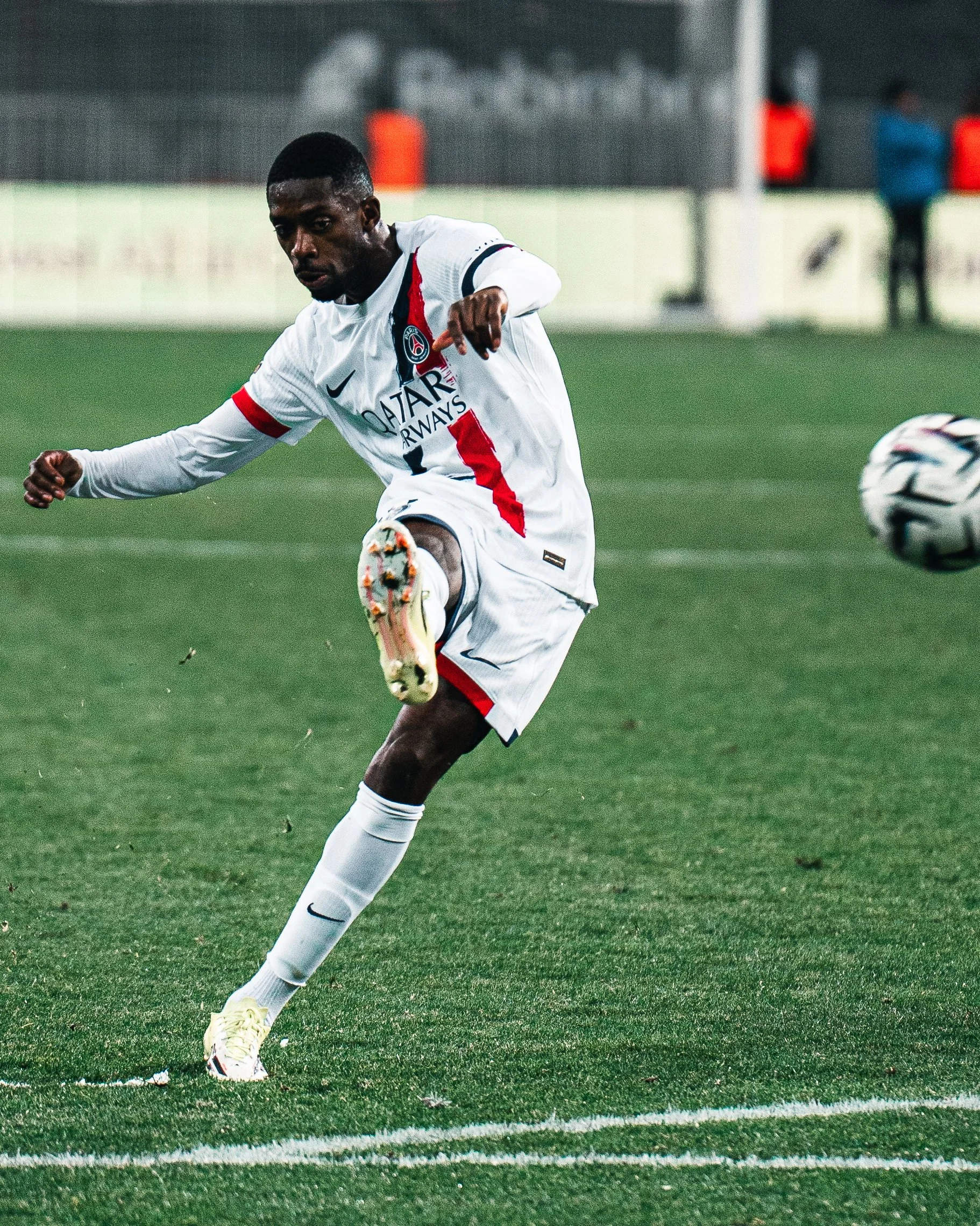 Un joueur de football en blanc en action lors d'un match, qui tente de frapper une balle sur un terrain vert, avec un fond flou de personnes et de structures.