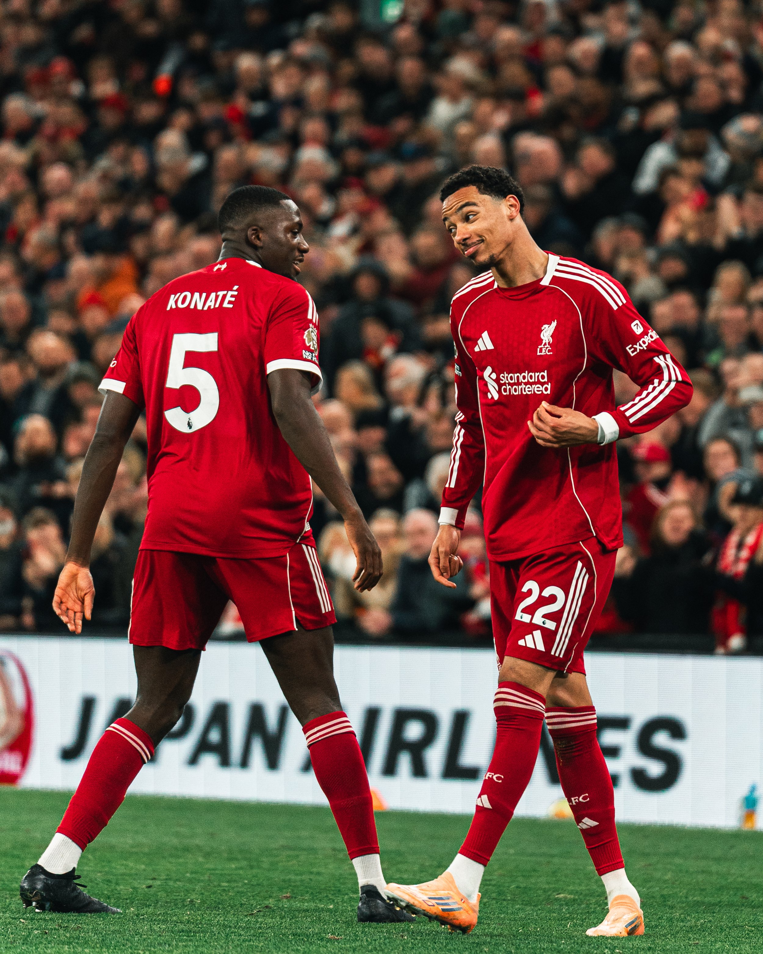 Deux joueurs du Liverpool FC en uniforme rouge discutent sur le terrain lors d'un match football, avec un public nombreux en arrière-plan.