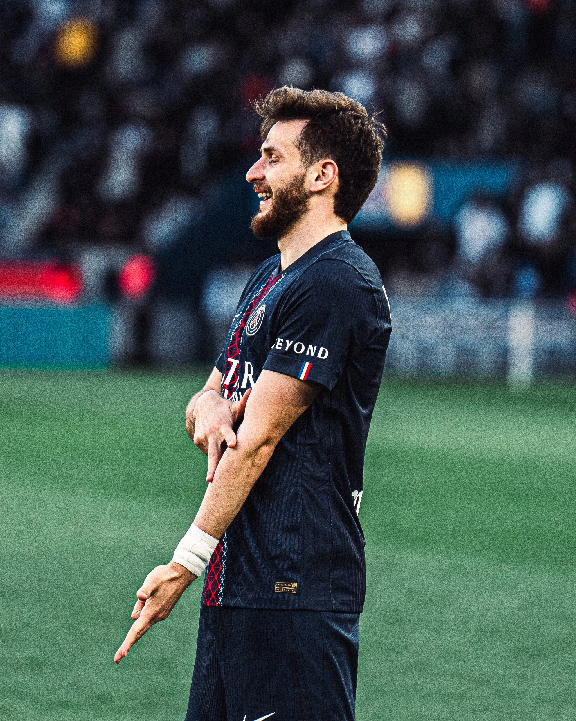 Un homme un sourire large, portant un maillot de football bleu, en train de célébrer un but ou un succès sur un terrain de football. Son bras est croisé et il porte une bande blanche sur le poignet gauche.