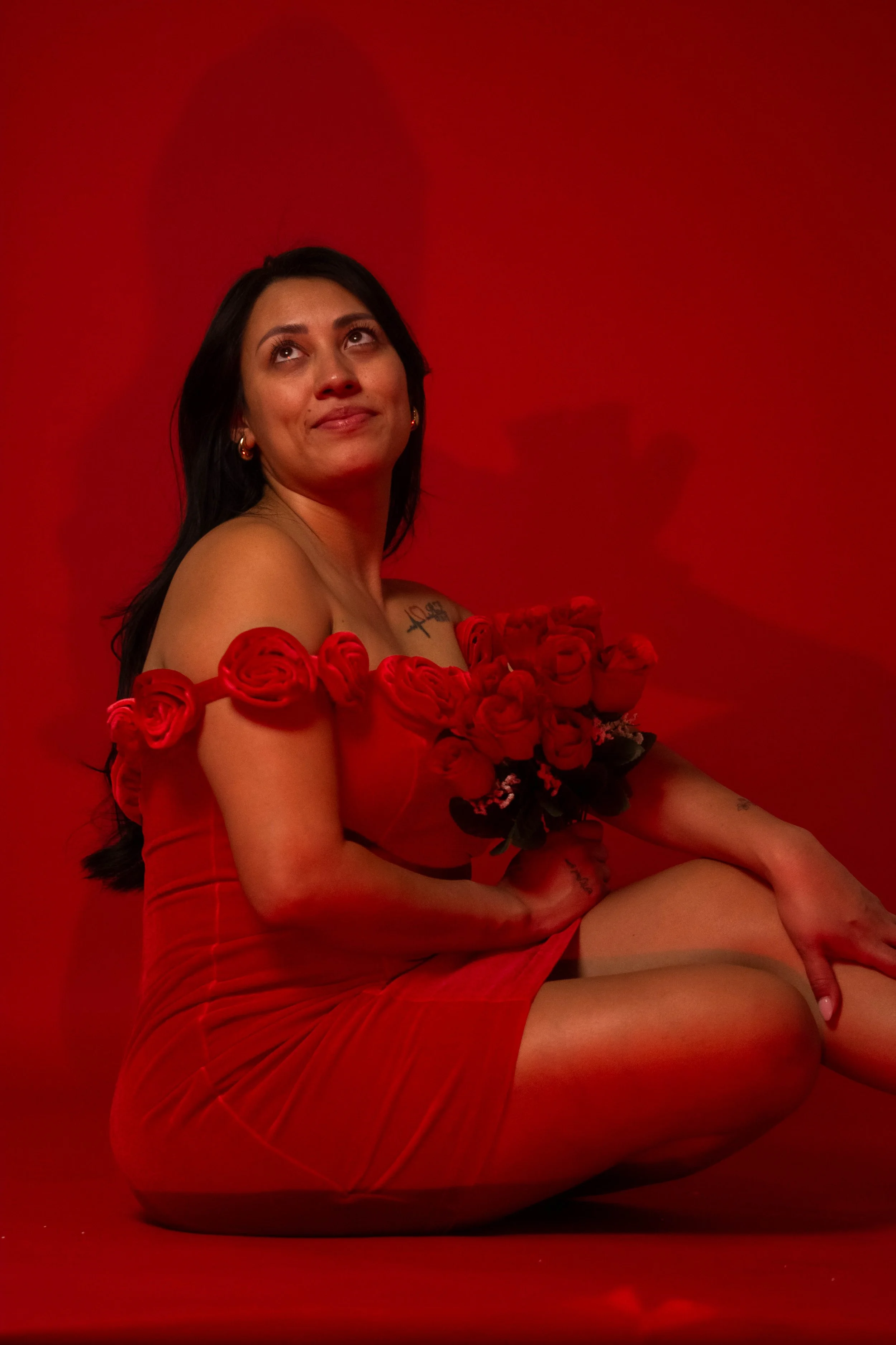 A woman sitting on the floor against a red background, holding a bouquet of red roses, wearing a red dress with rose details on the shoulders, looking up with a slight smile.