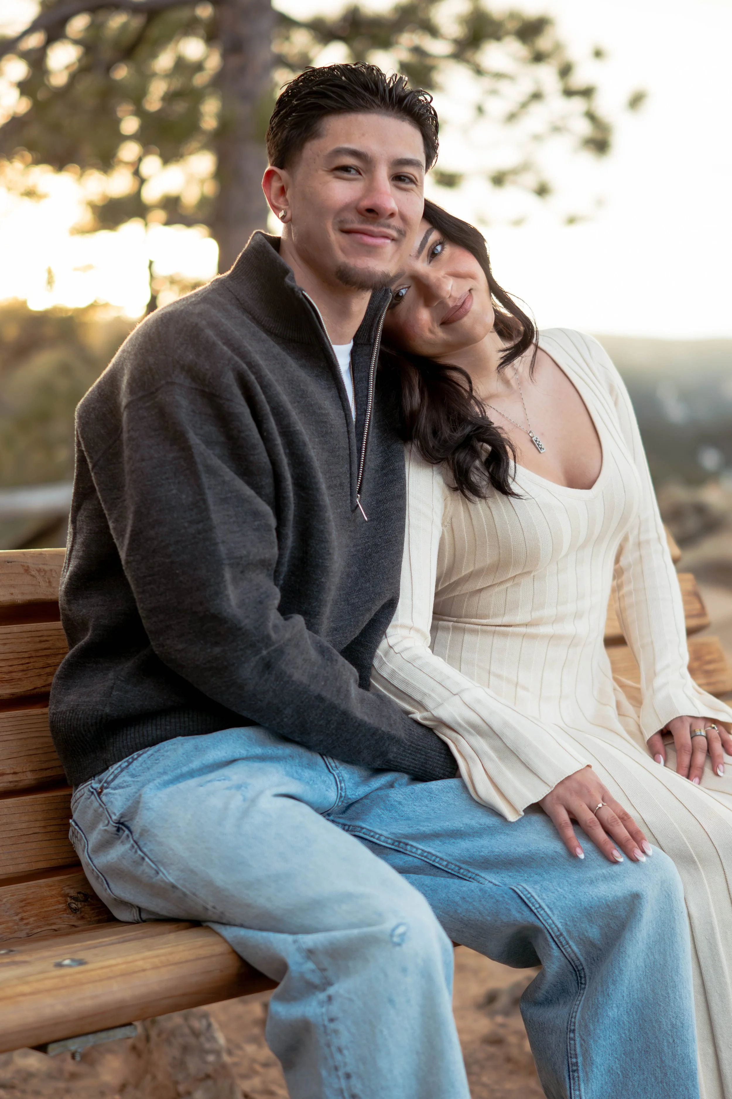 A young couple sitting on a wooden bench outdoors, leaning against each other, with trees and sunset in the background.