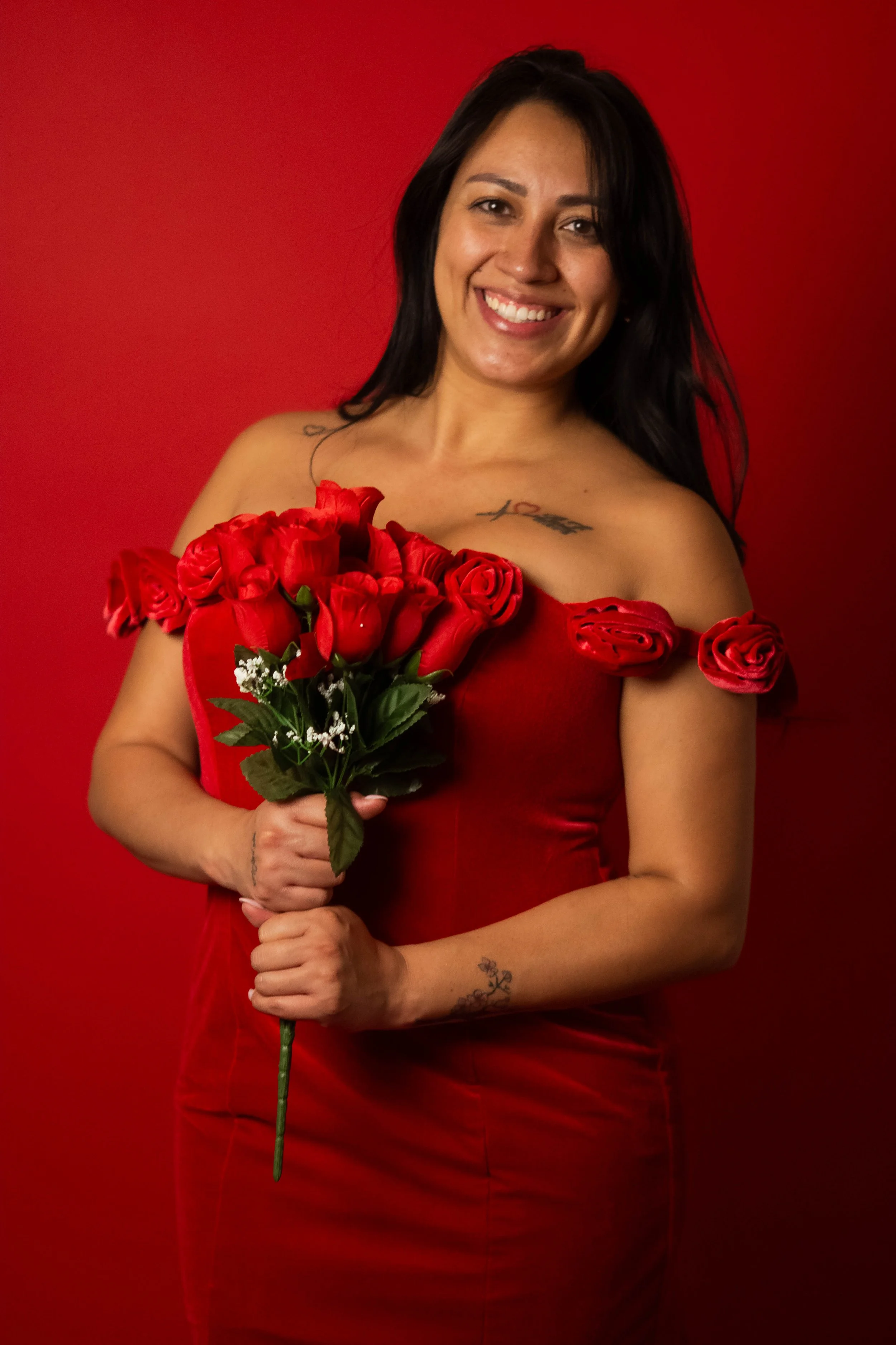 A woman dressed in a red off-the-shoulder dress with roses on the shoulders, holding a bouquet of red roses, smiling in front of a red background.