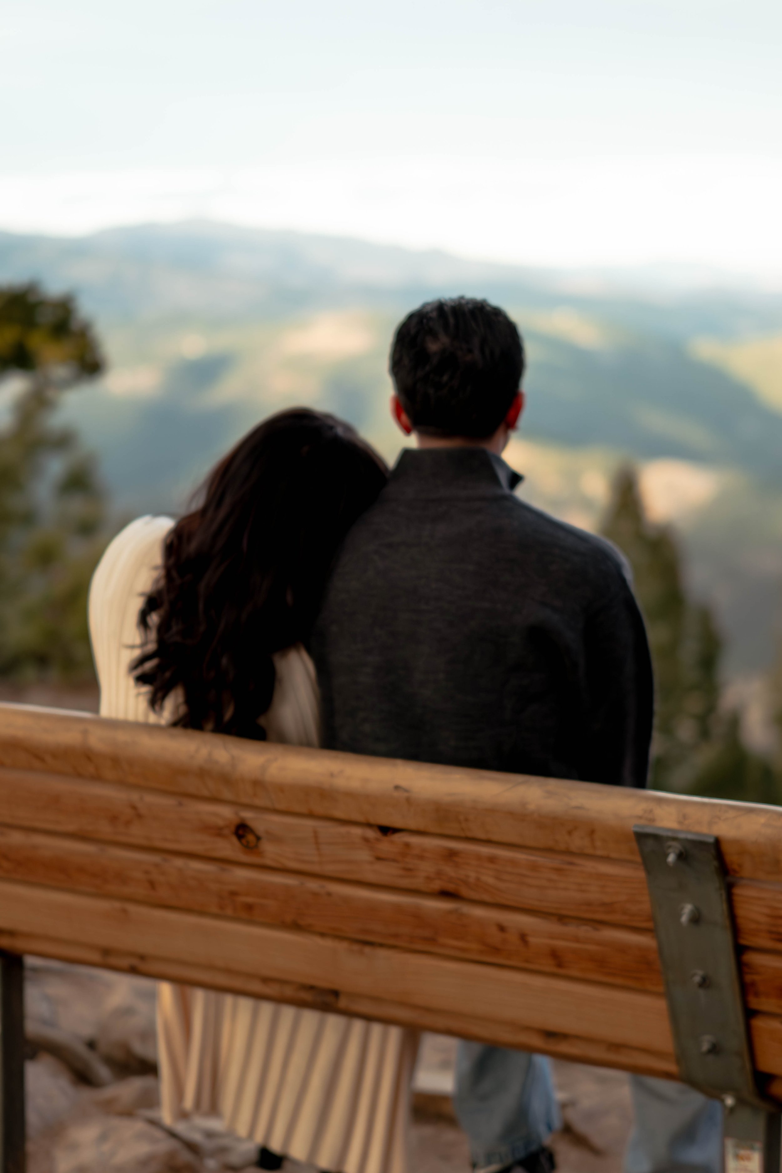 A man and woman sitting close together on a bench, looking at a scenic view of hills and trees.