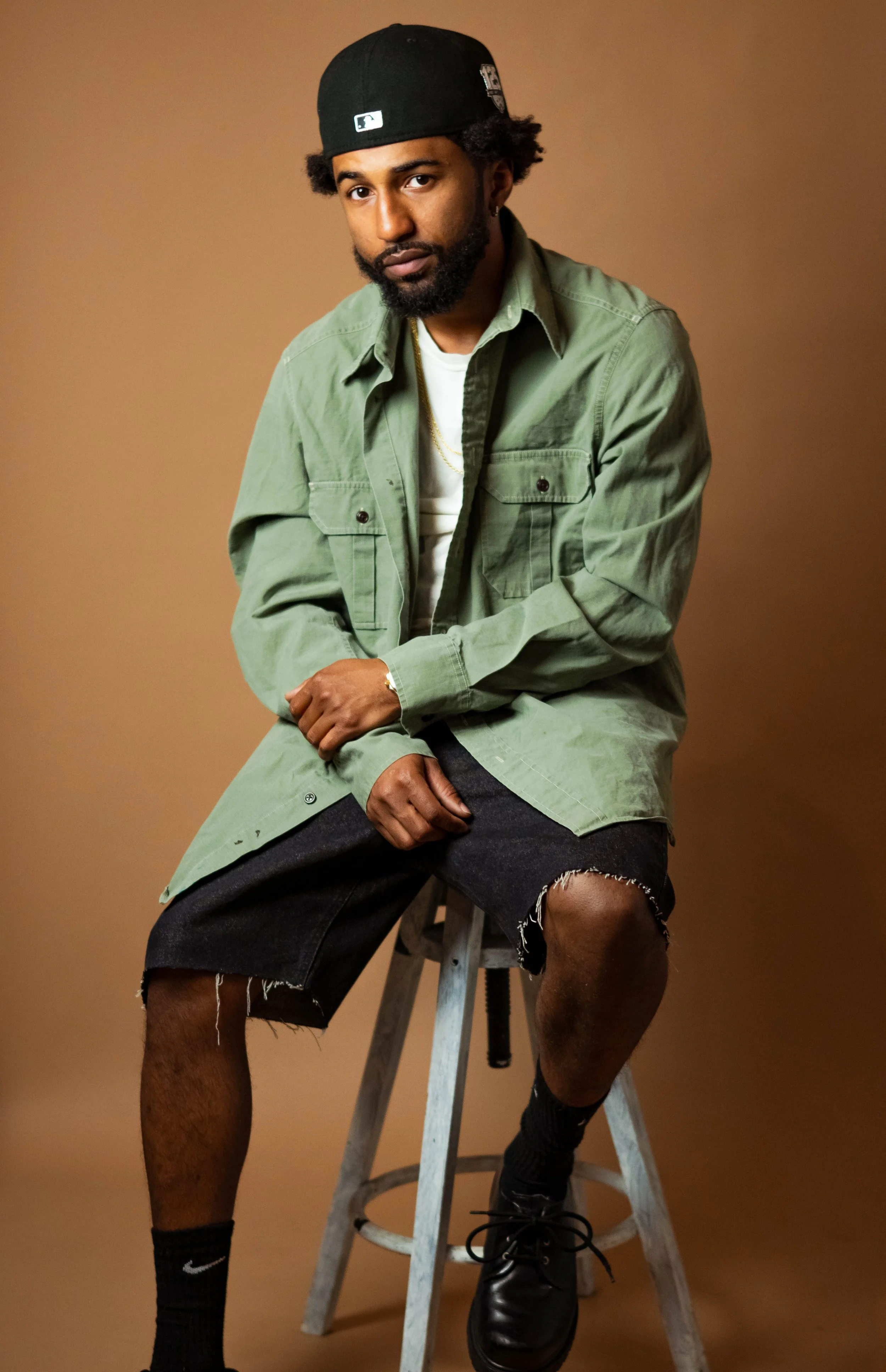 A young man with a beard and dark curly hair wearing a black baseball cap, green overshirt, white t-shirt, ripped black shorts, black socks, and black shoes, sitting on a wooden stool against a brown background.