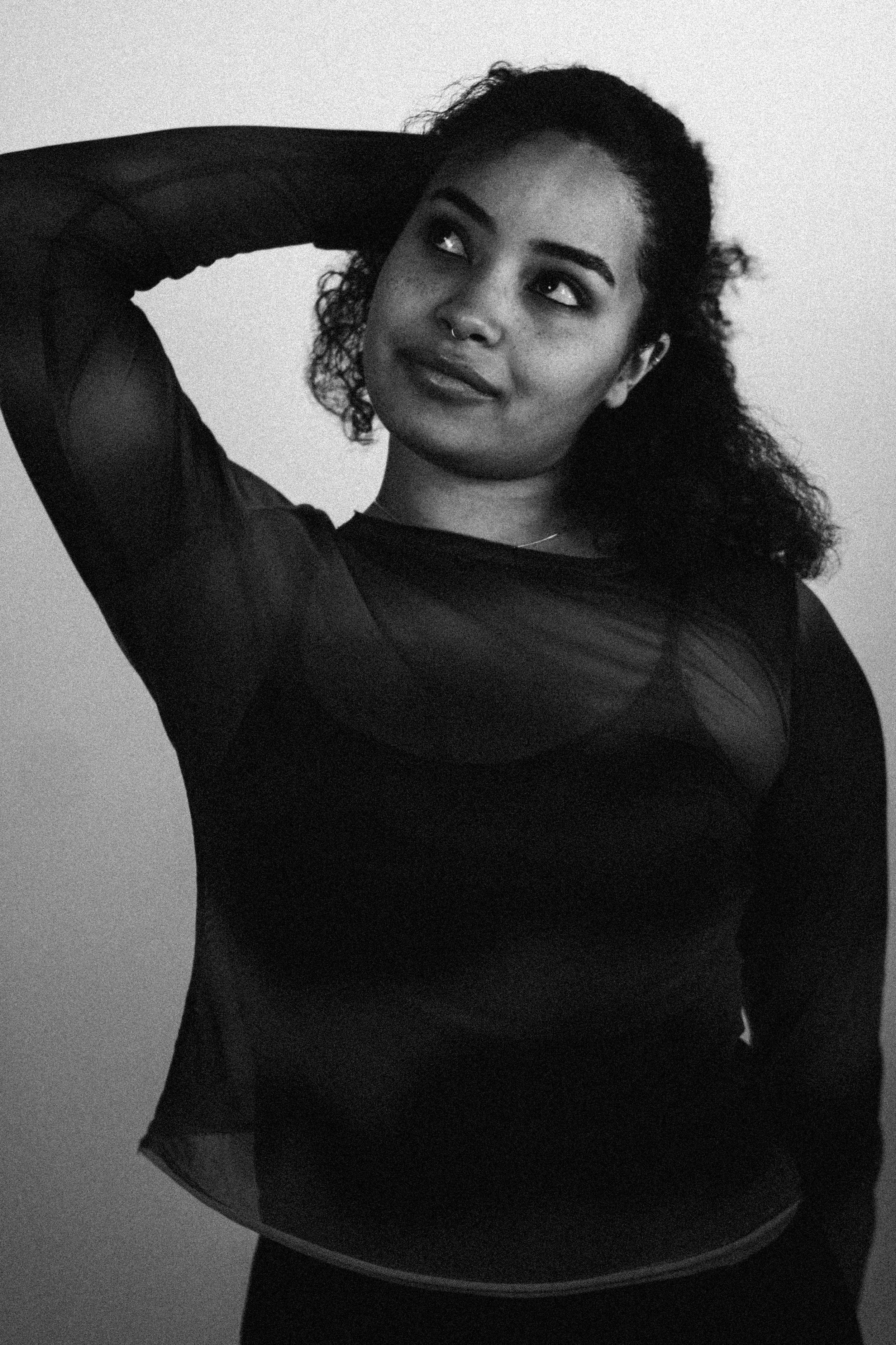 Black and white portrait of a young woman with curly hair, wearing a sheer black top, raising one arm behind her head, looking to the side with a slight smile.