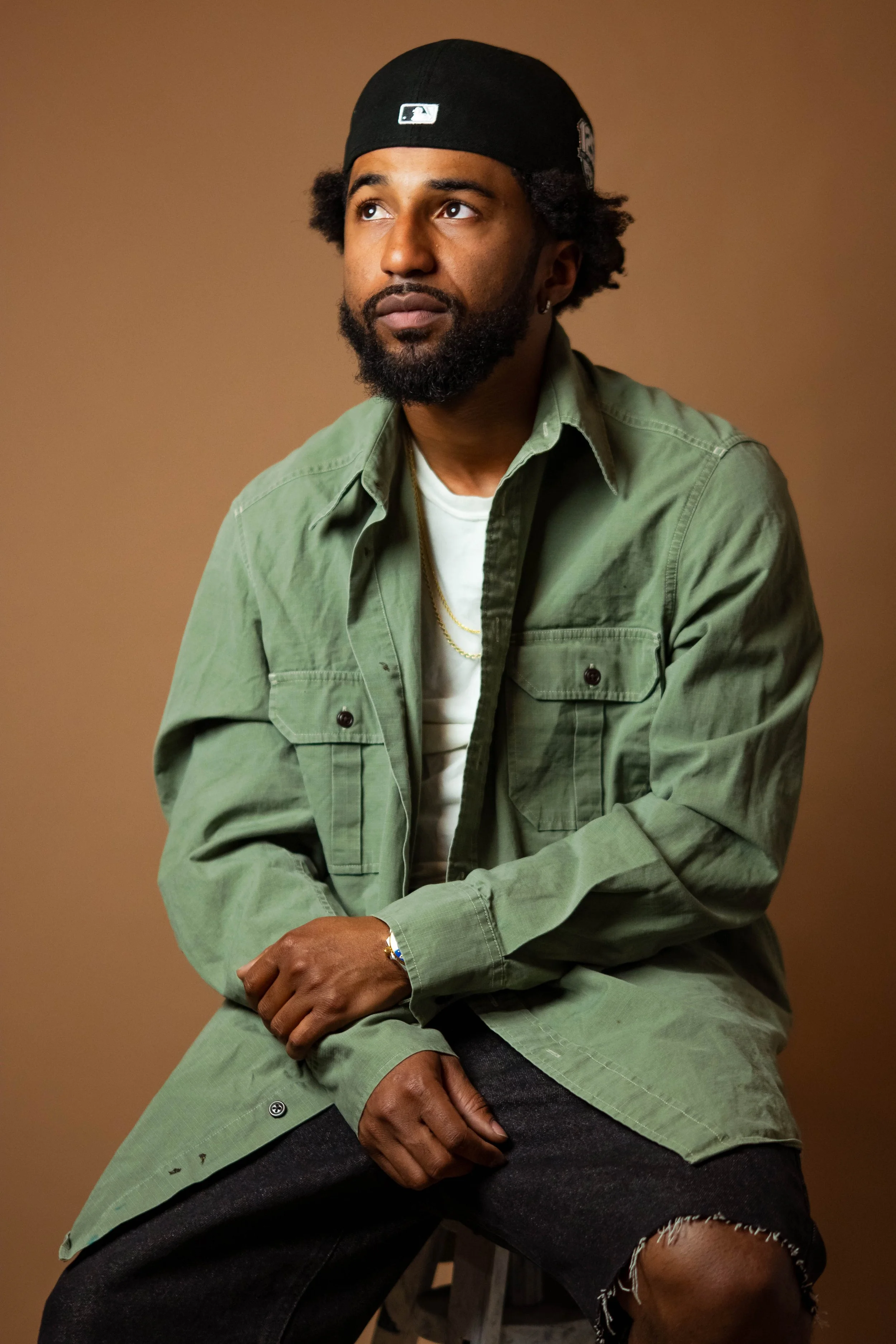A man with dark skin, beard, and curly hair wearing a black cap, green jacket, white t-shirt, black shorts, and jewelry, seated against a plain brown background.