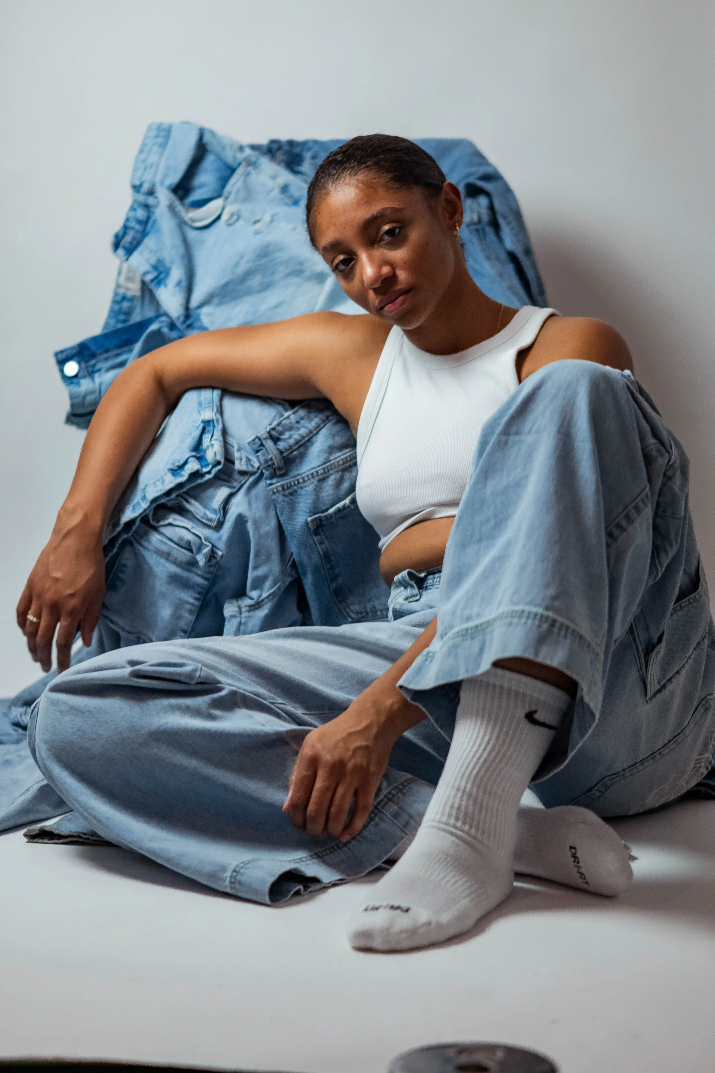 A young woman in a white sleeveless top and jeans, with a white Nike sock showing, sitting on the floor in front of a pile of denim clothes. She has short hair, gold earrings, and a subtle expression.