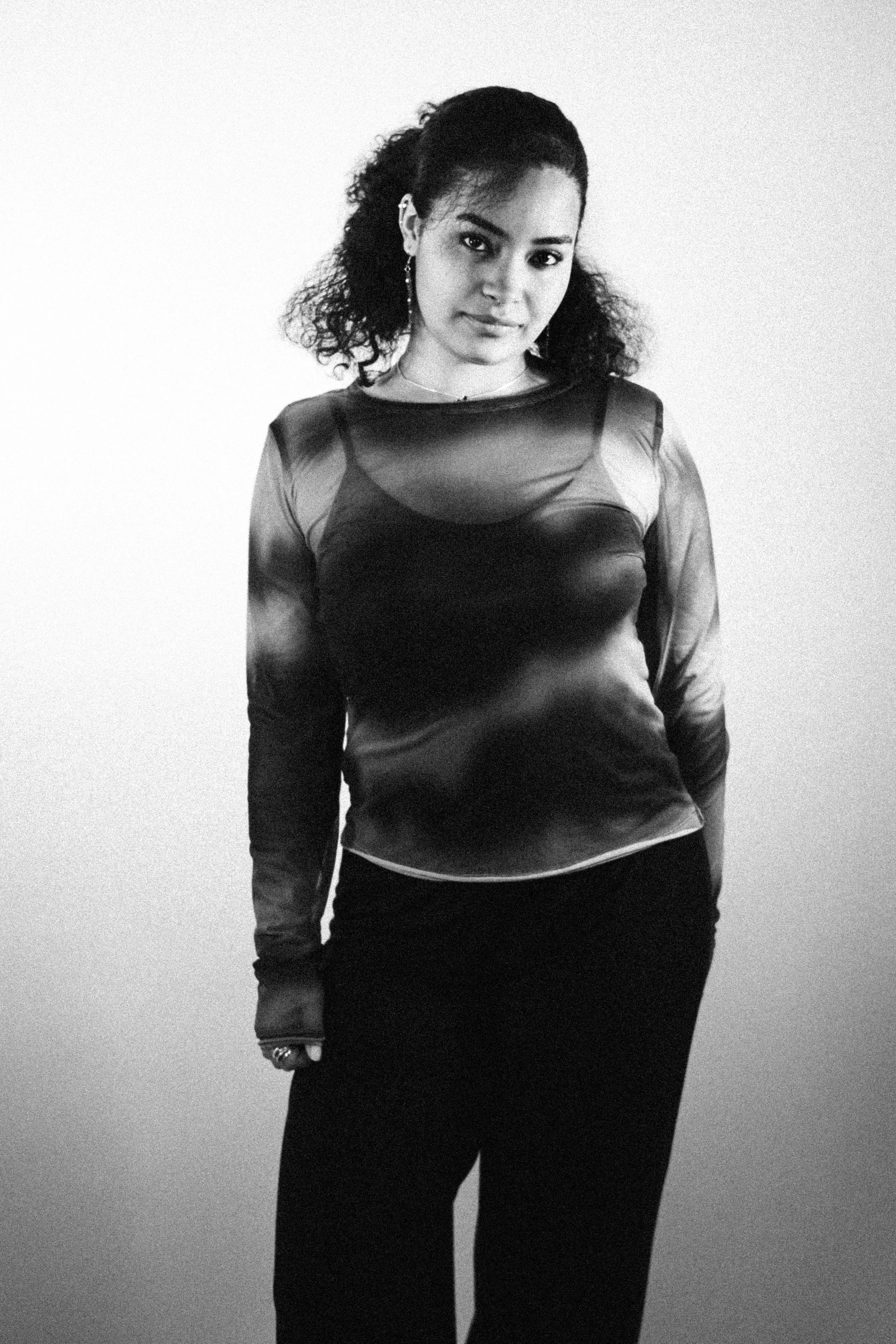 A young woman with curly hair, wearing a sheer, long-sleeved top and dark pants, standing against a plain wall.