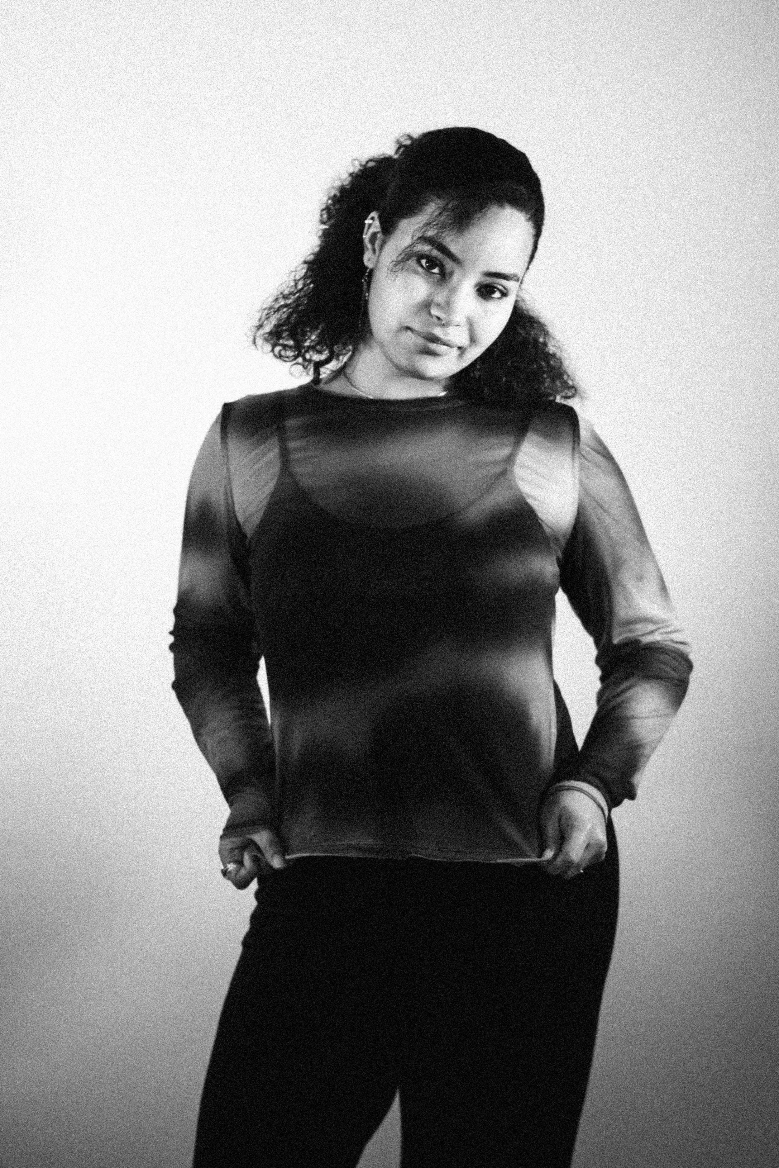 A woman with curly hair in a black sheer top, posing against a plain wall.