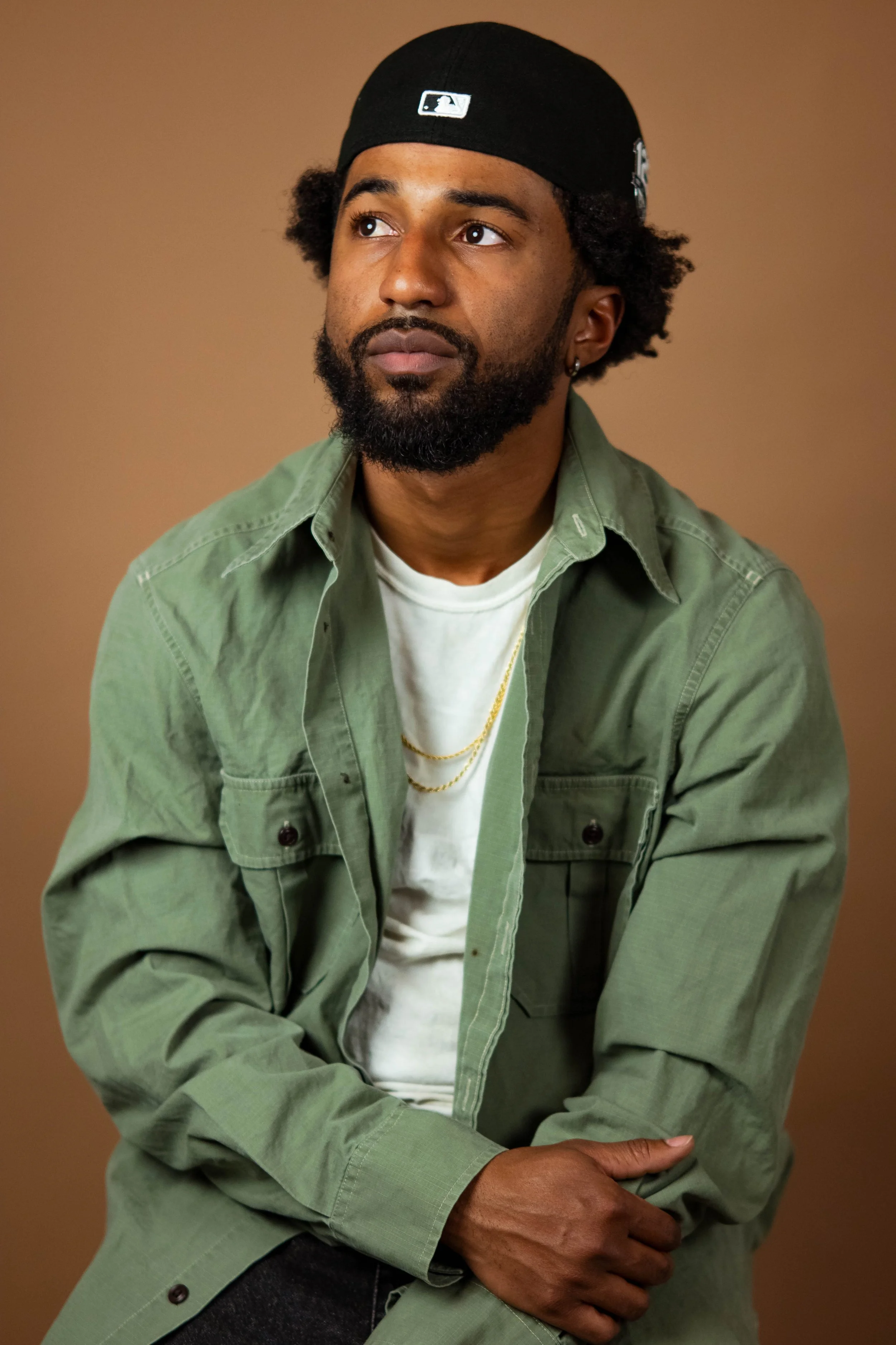 Portrait of a bearded Black man with curly hair, wearing a black baseball cap, green jacket, white T-shirt, and gold chains, seated against a brown background.