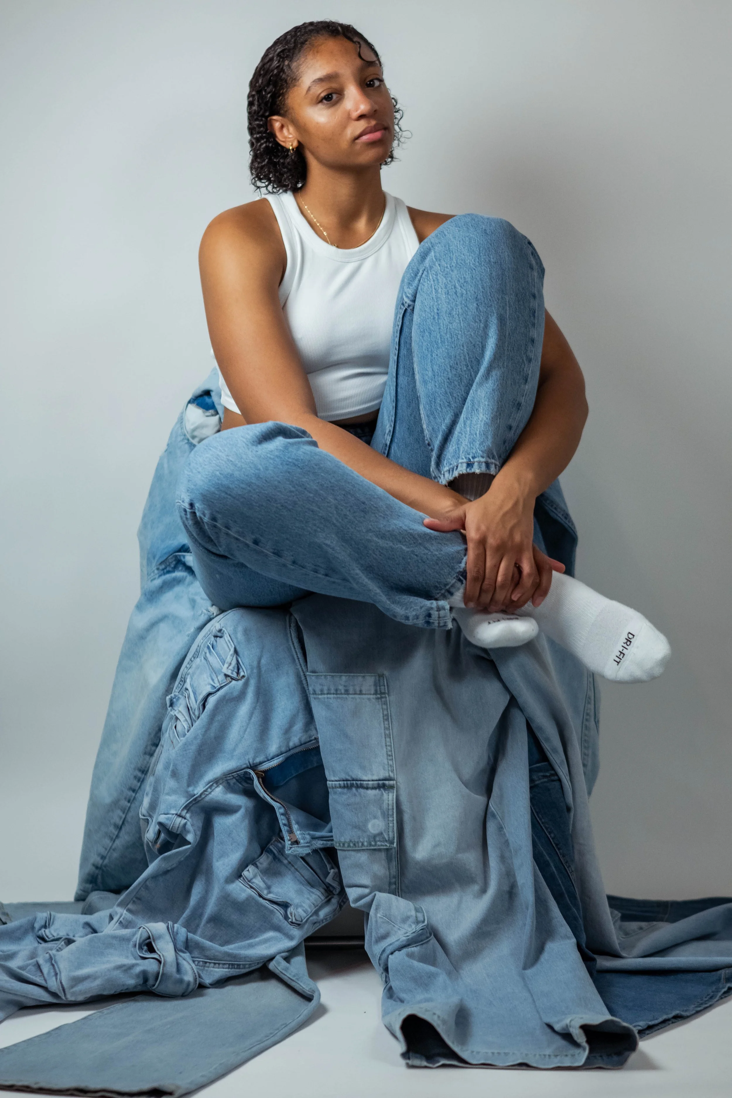 A young woman with short curly hair and a tank top sitting on a large pile of blue jeans and denim clothing, with a plain light gray background.