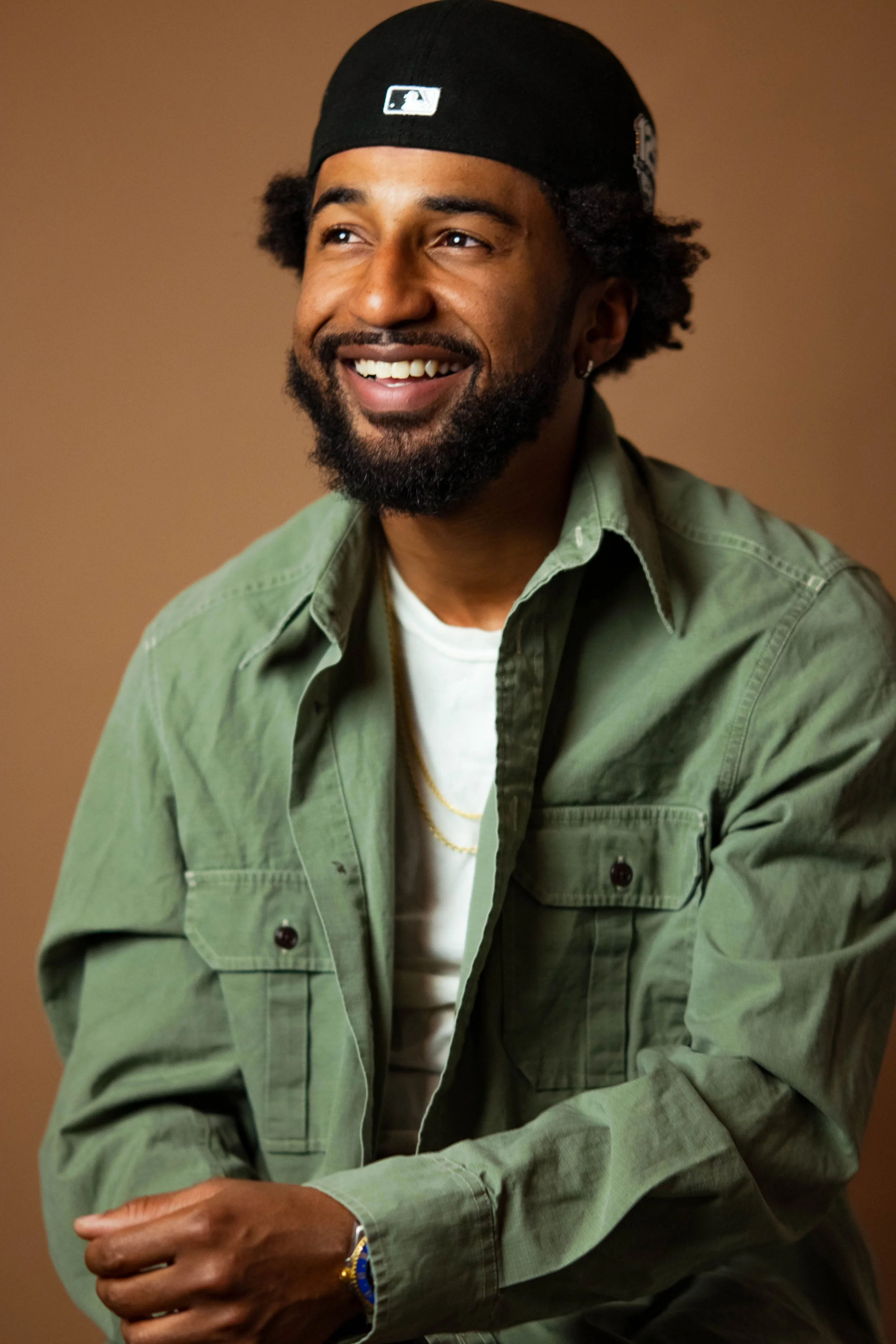 A man with curly hair and a beard, smiling, wearing a black cap, an olive-green jacket over a white T-shirt, and gold jewelry, against a plain brown background.