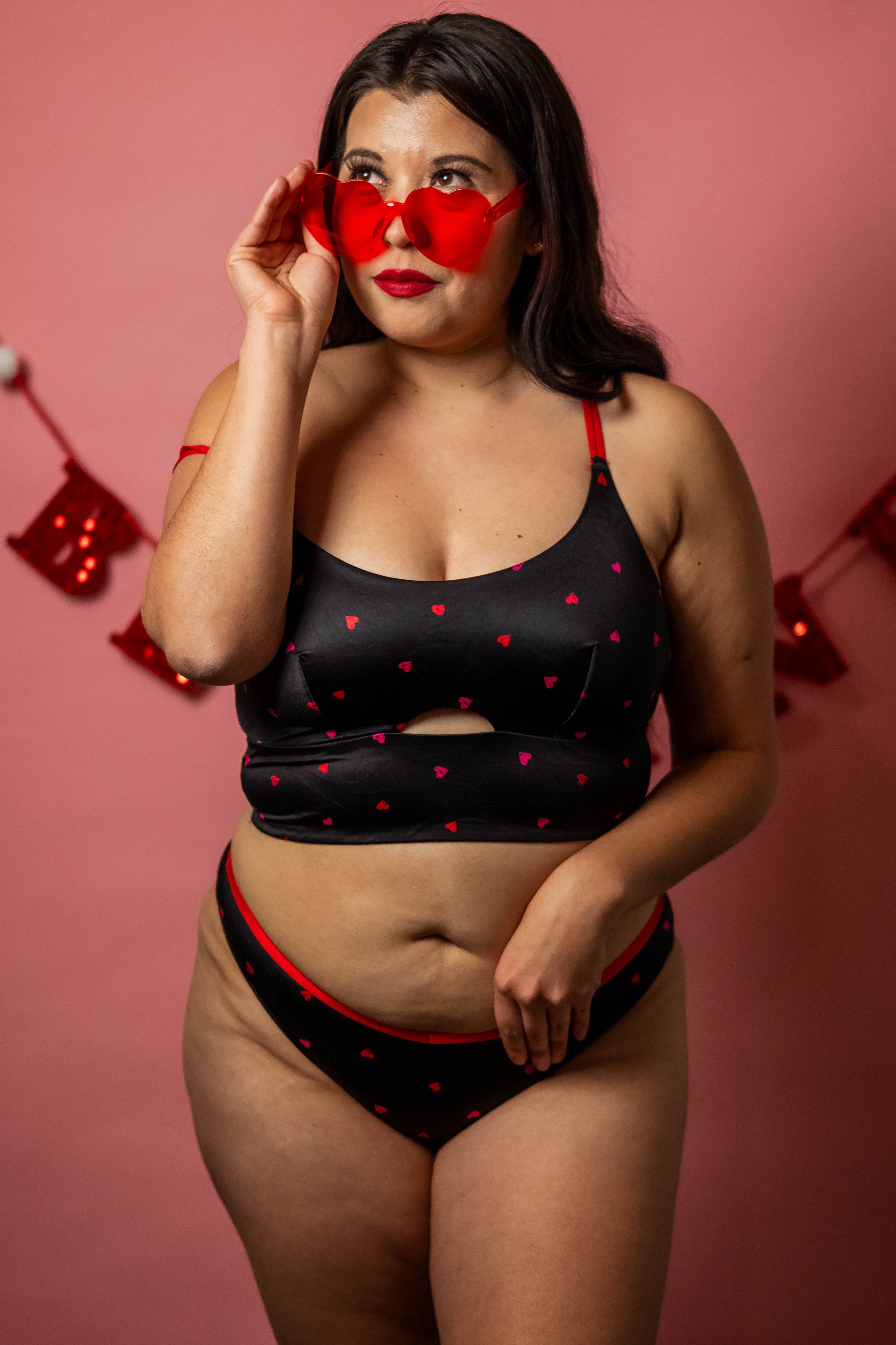 Plus-size woman wearing black lingerie with red hearts, red heart-shaped sunglasses, standing against pink wall decorated with red banners.