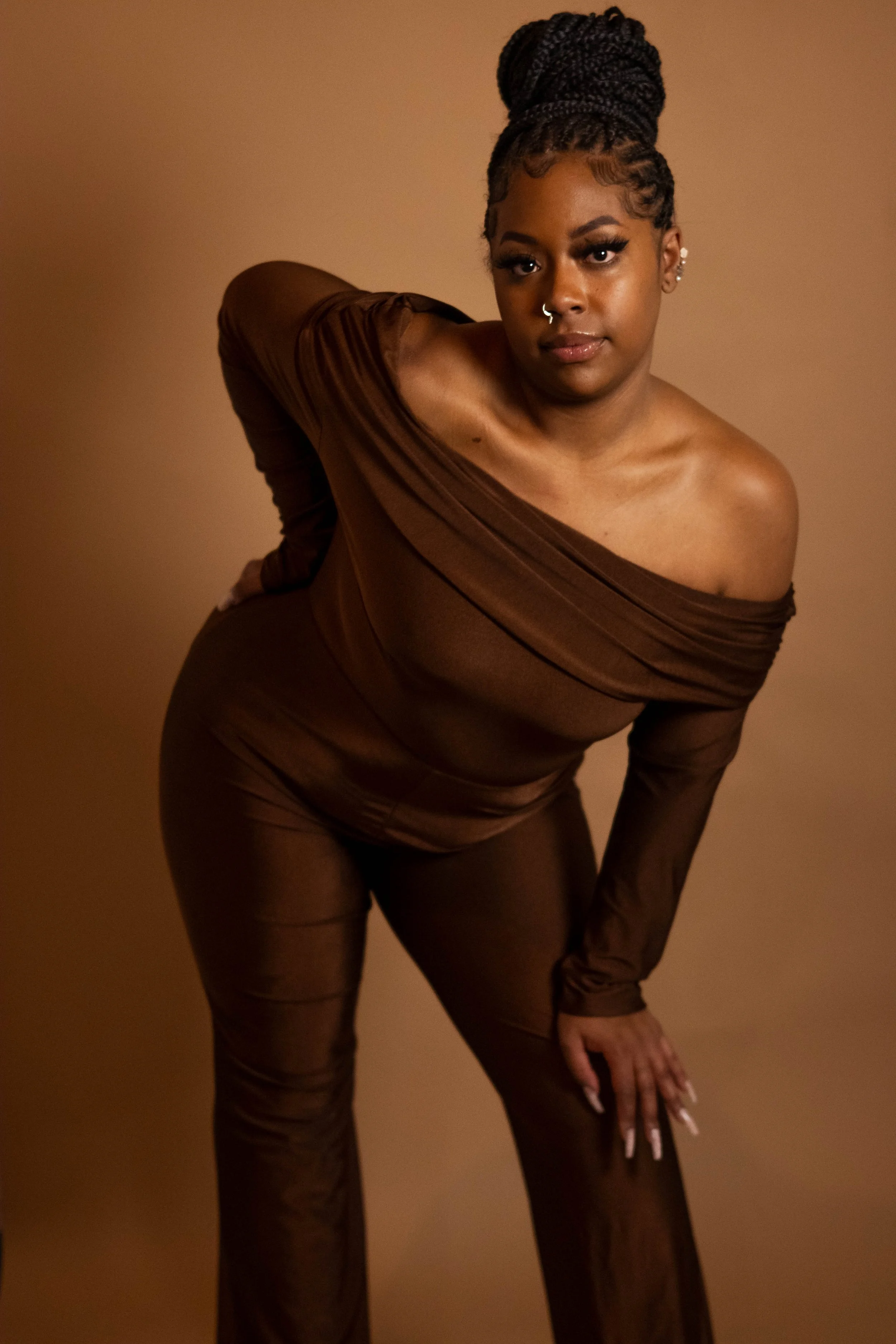 A woman with braided hair styled in a high bun, wearing a sheer brown off-the-shoulder top and matching pants, posed against a plain brown background.
