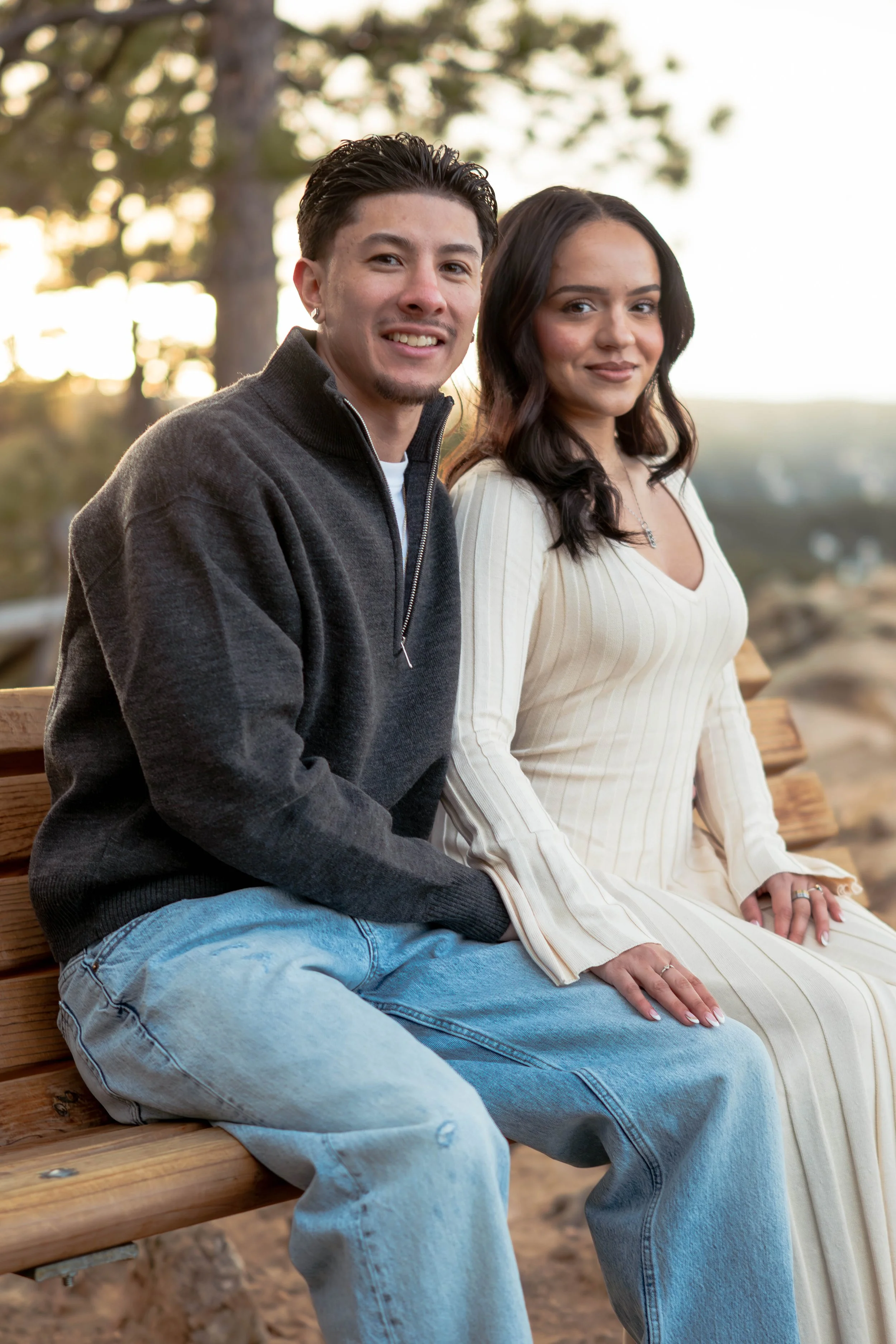 A young man and woman sitting on a wooden bench outdoors, smiling, with trees and a sunset in the background.