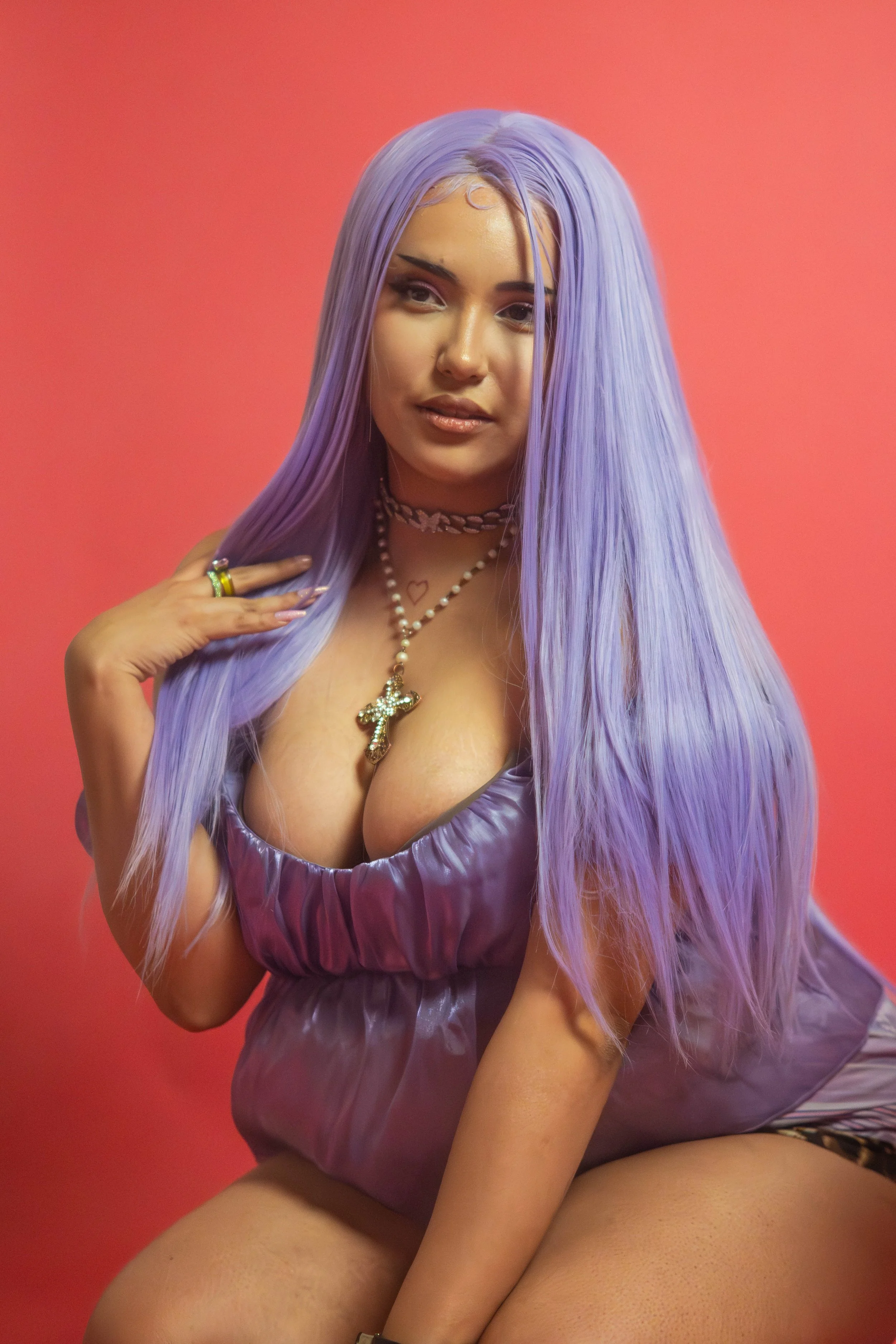 A woman with long, lavender hair posing with a pink background, wearing a purple satin top, layered necklaces including a cross pendant, and rings, with makeup and glossy lips.
