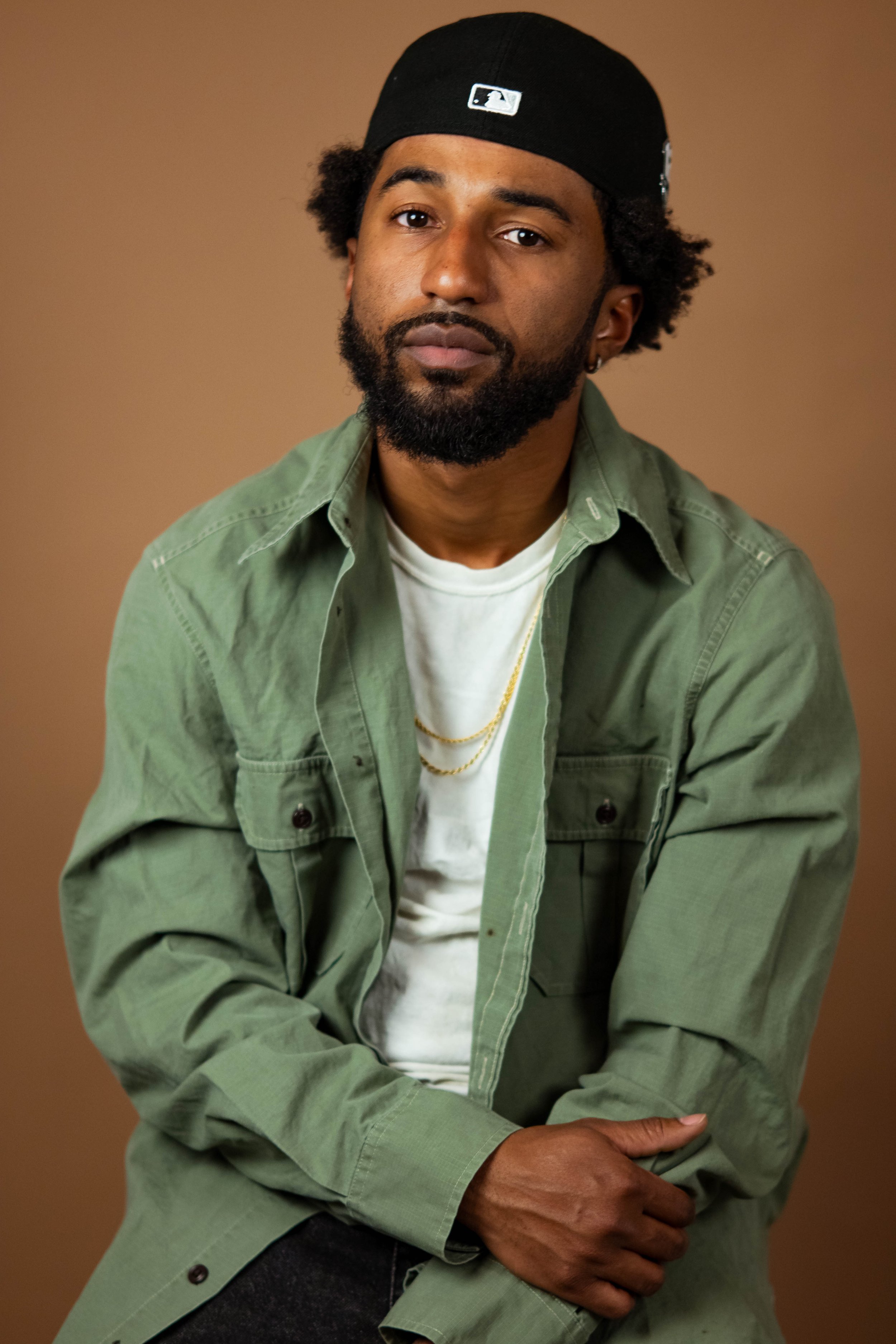 Portrait of a man with a beard and curly hair wearing a black cap, green jacket, white T-shirt, and gold chains, sitting against a plain brown background.