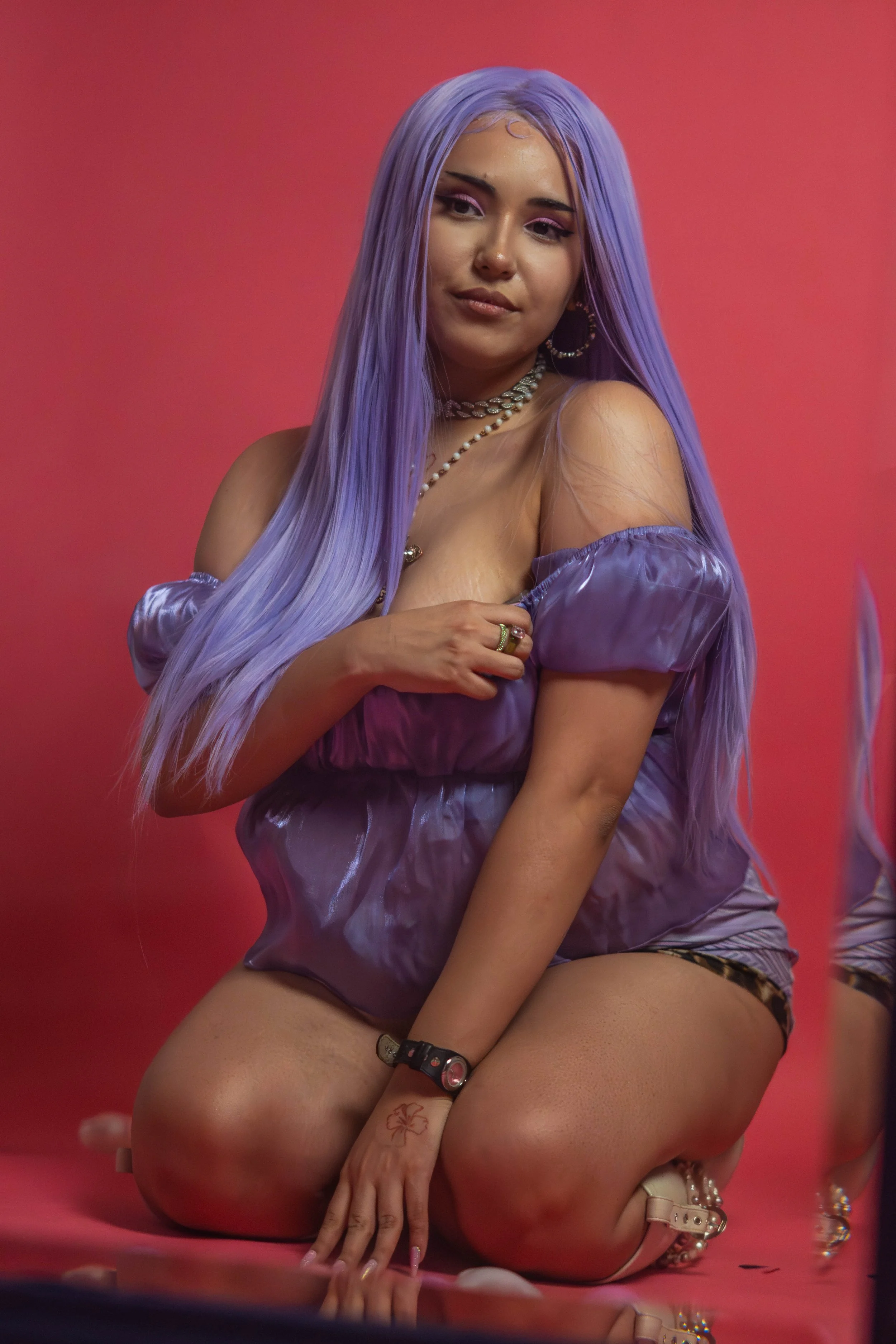 Woman with long purple hair kneeling on pink floor against pink background, wearing purple satin off-shoulder dress, jewelry, and shoes with pearl details.
