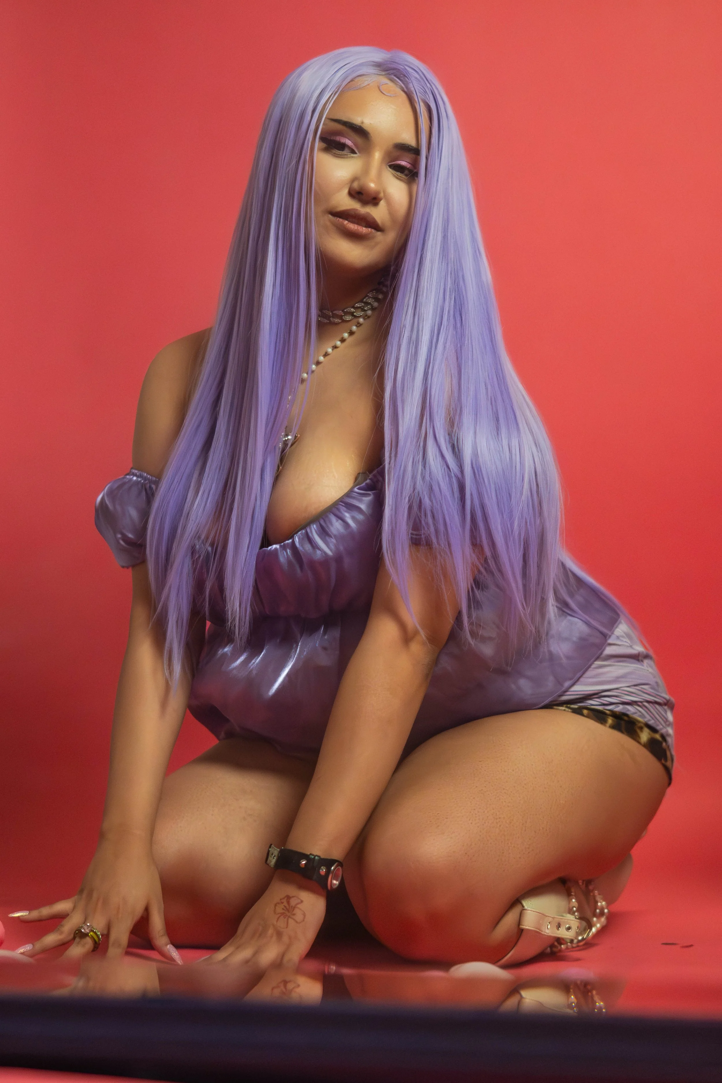 A woman with long purple hair kneeling on the floor with a pink background, wearing a shiny purple dress, jewelry, and animal print shorts.