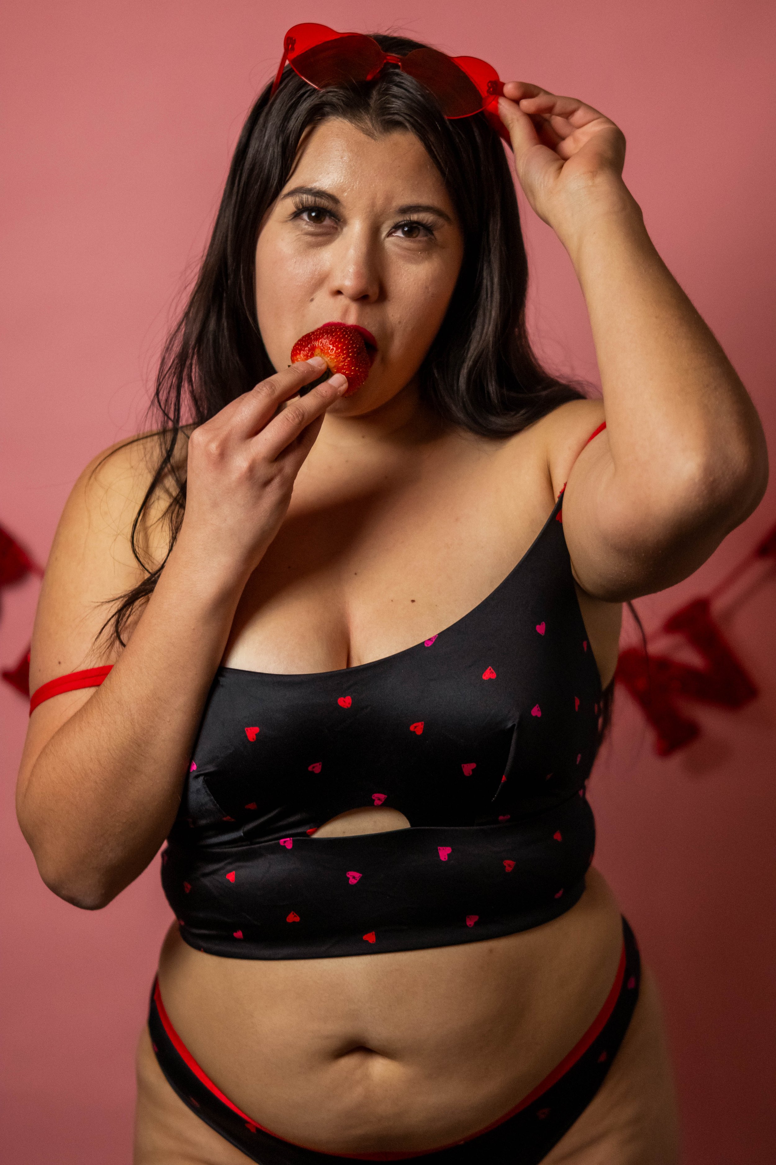 A woman with long dark hair wearing heart-patterned black lingerie with red accents, holding a strawberry near her mouth and adjusting red sunglasses on her head, standing against a pink background with red decorations.