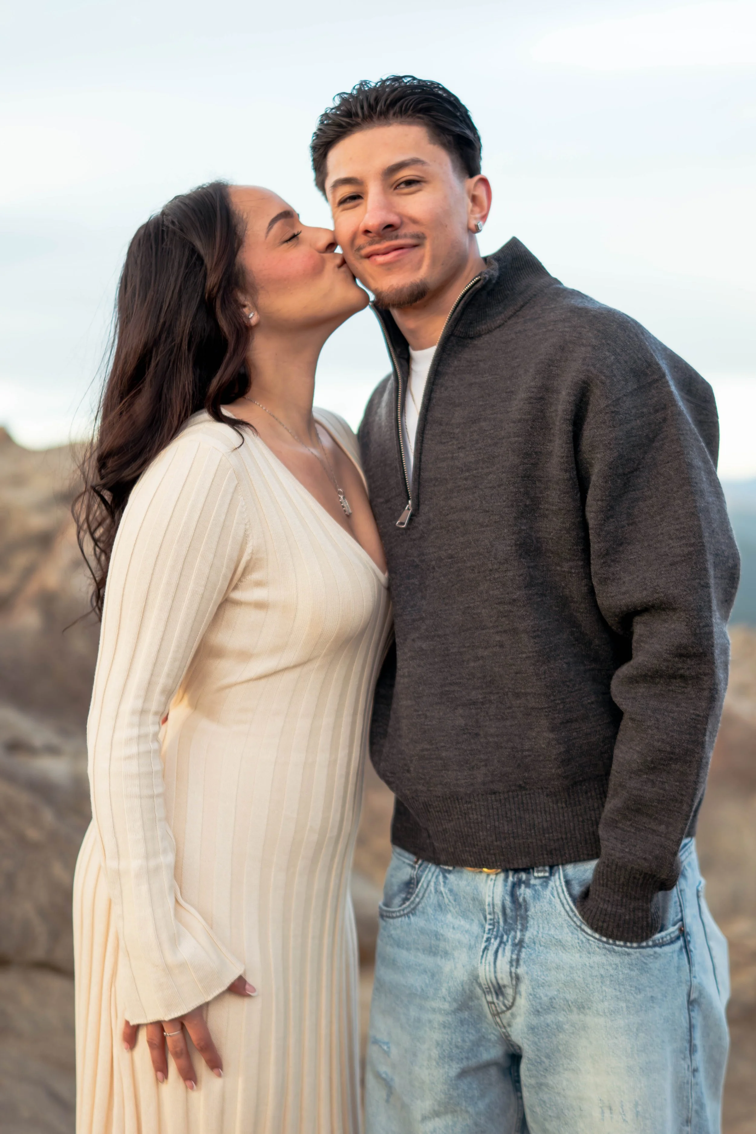 A woman is kissing a man on the cheek in an outdoor setting with rocky terrain, and the man is smiling at the camera.