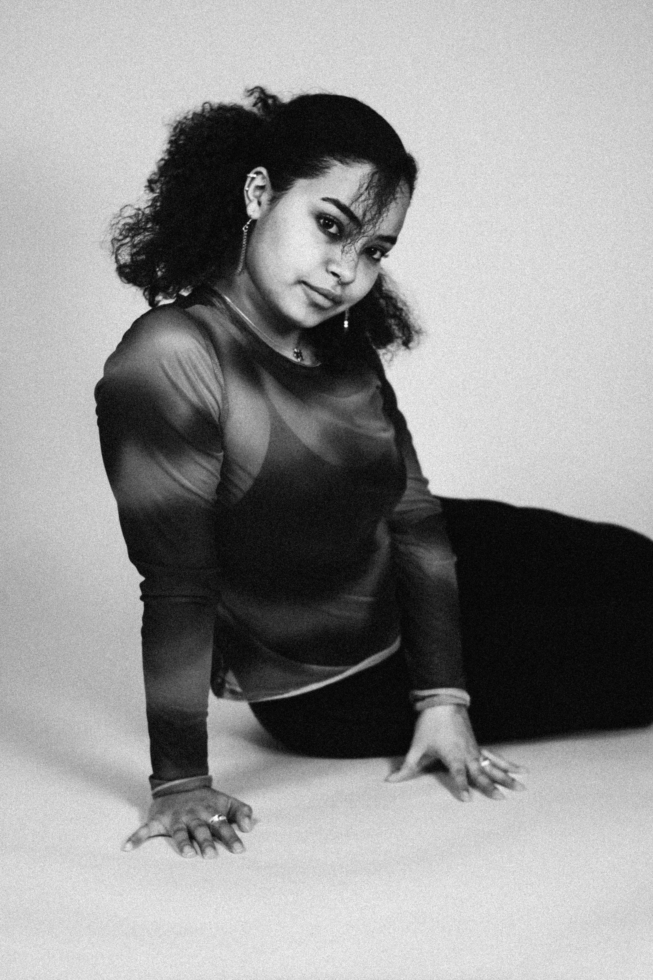 A young woman with curly hair, dressed in a sheer long-sleeve top, sitting on the ground with her hands on the floor in front of her, looking at the camera.