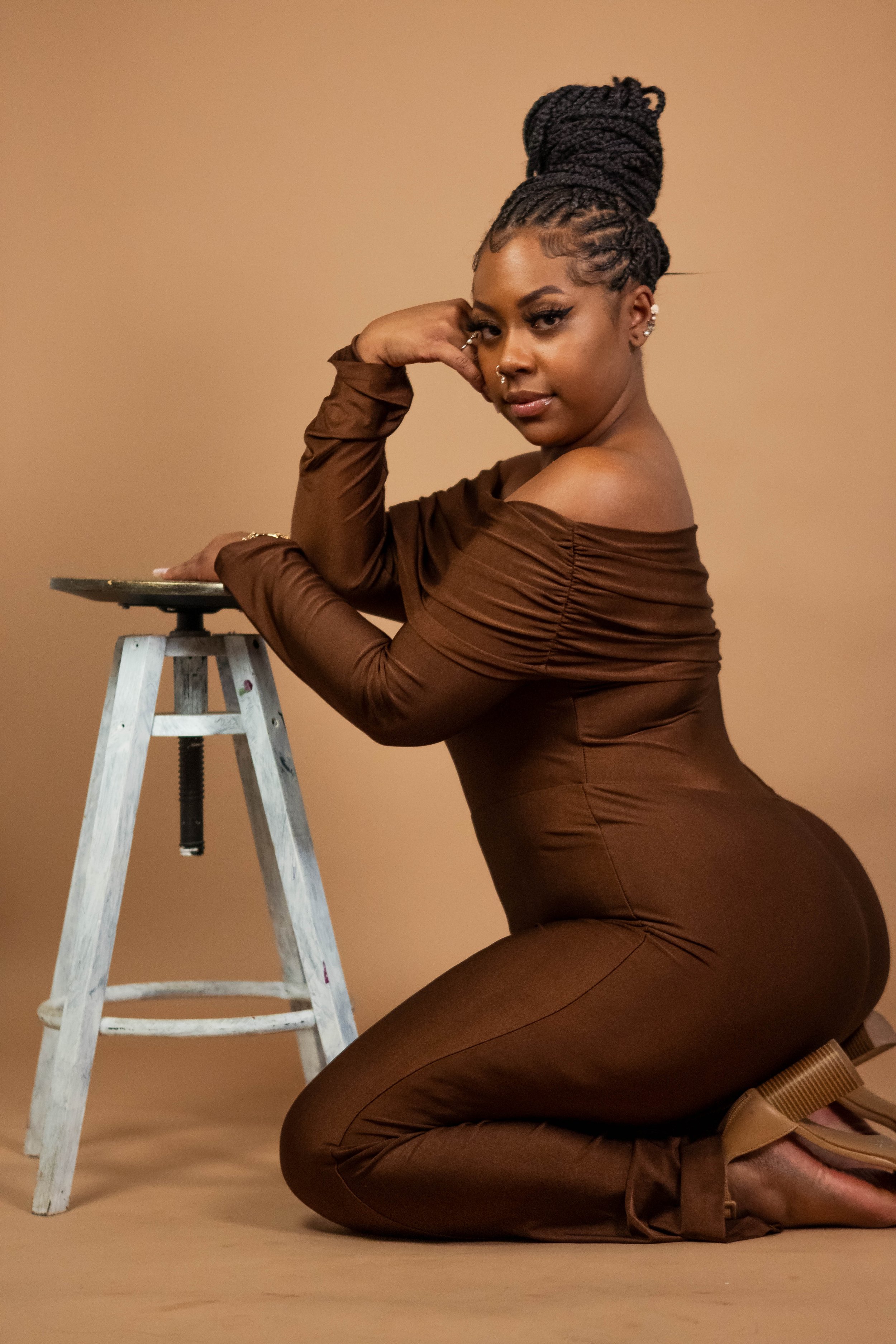 A woman in a brown off-shoulder dress kneels on the floor, resting her left arm on a small step ladder while looking at the camera against a beige background.