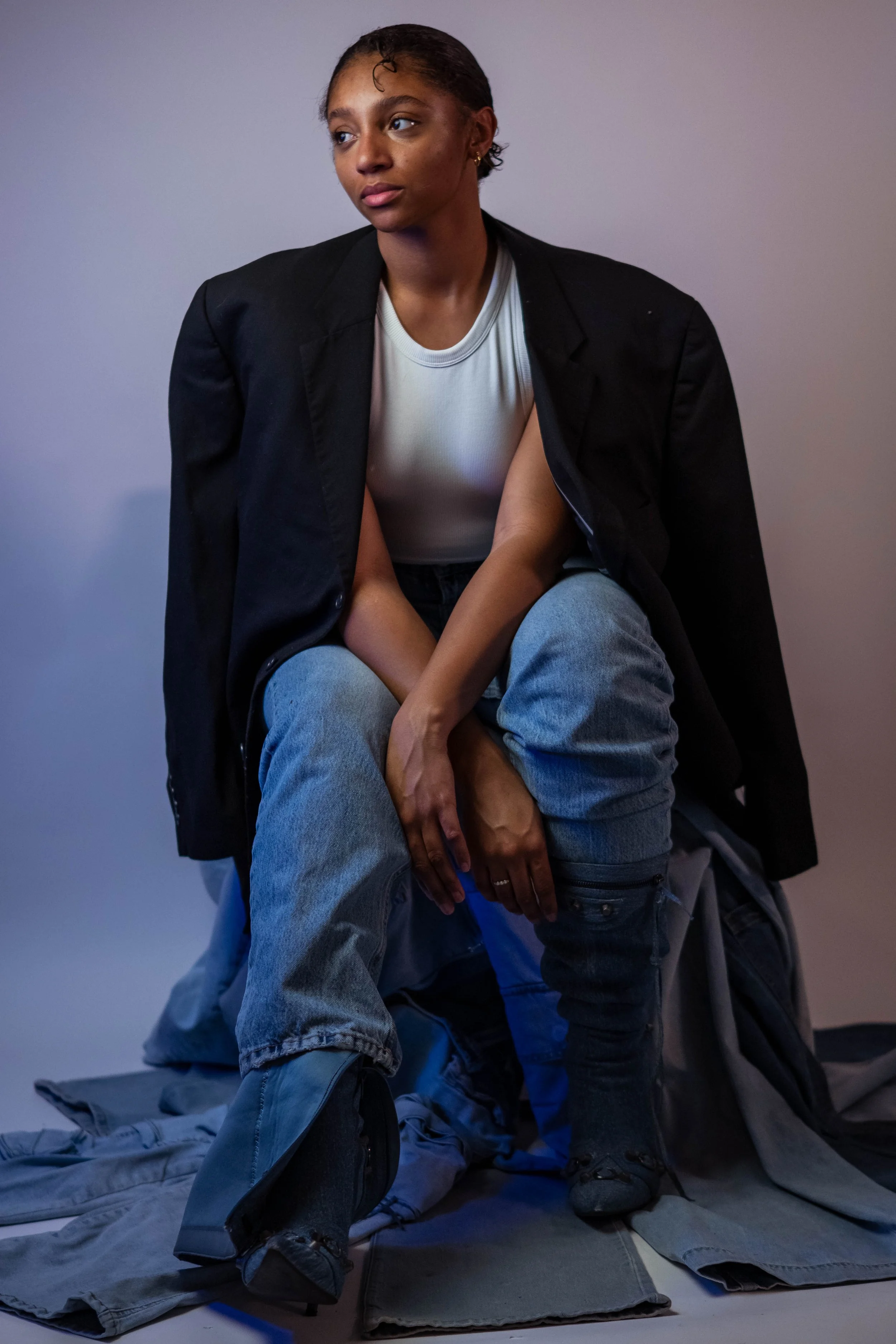 A young woman with natural hair, wearing a white t-shirt, black blazer, and jeans, sitting on a pile of clothes against a plain background.