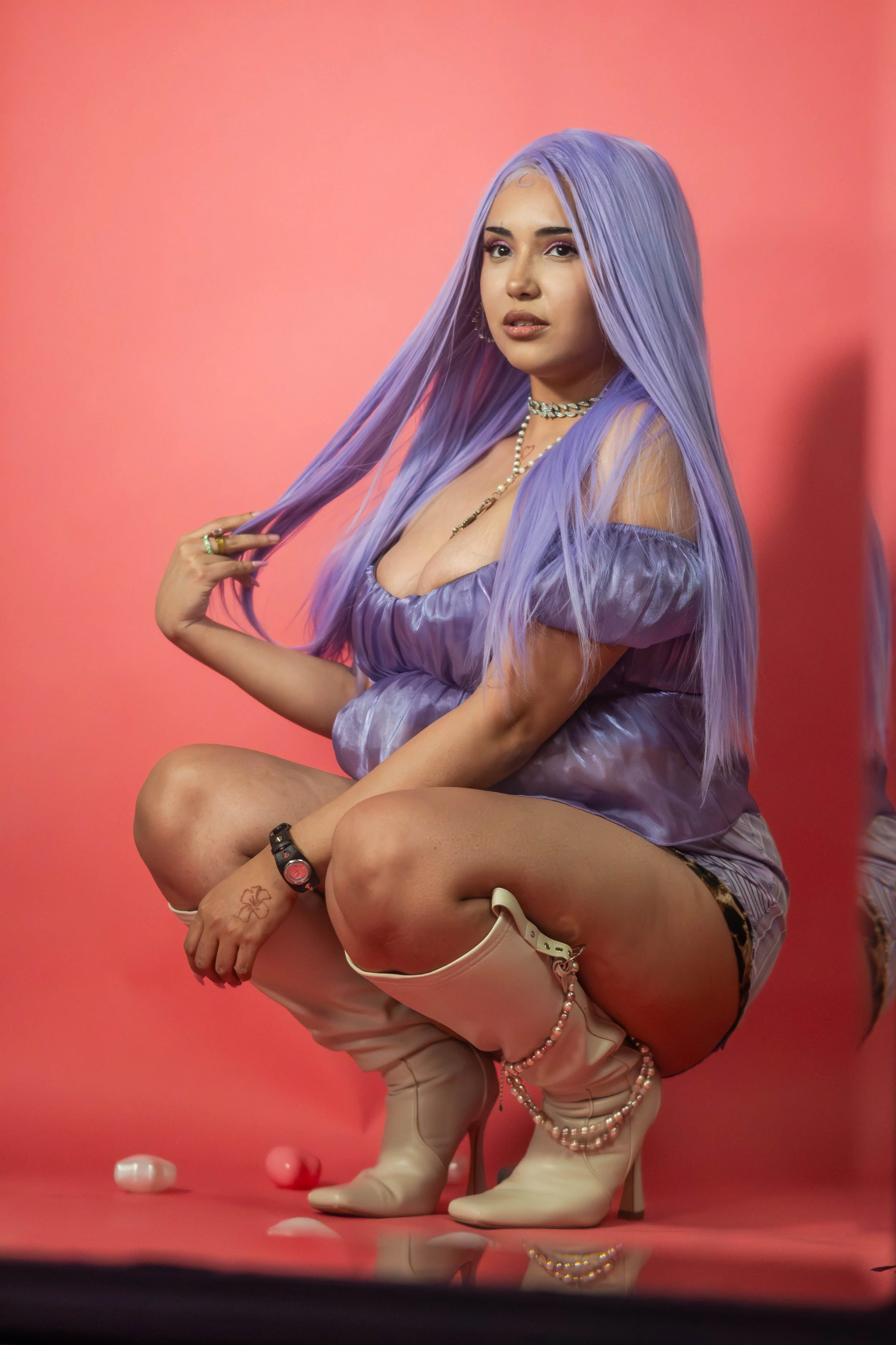 A woman with long purple hair squatting, wearing a purple off-shoulder top, leopard print shorts, and white high-heeled boots, with a pink background.