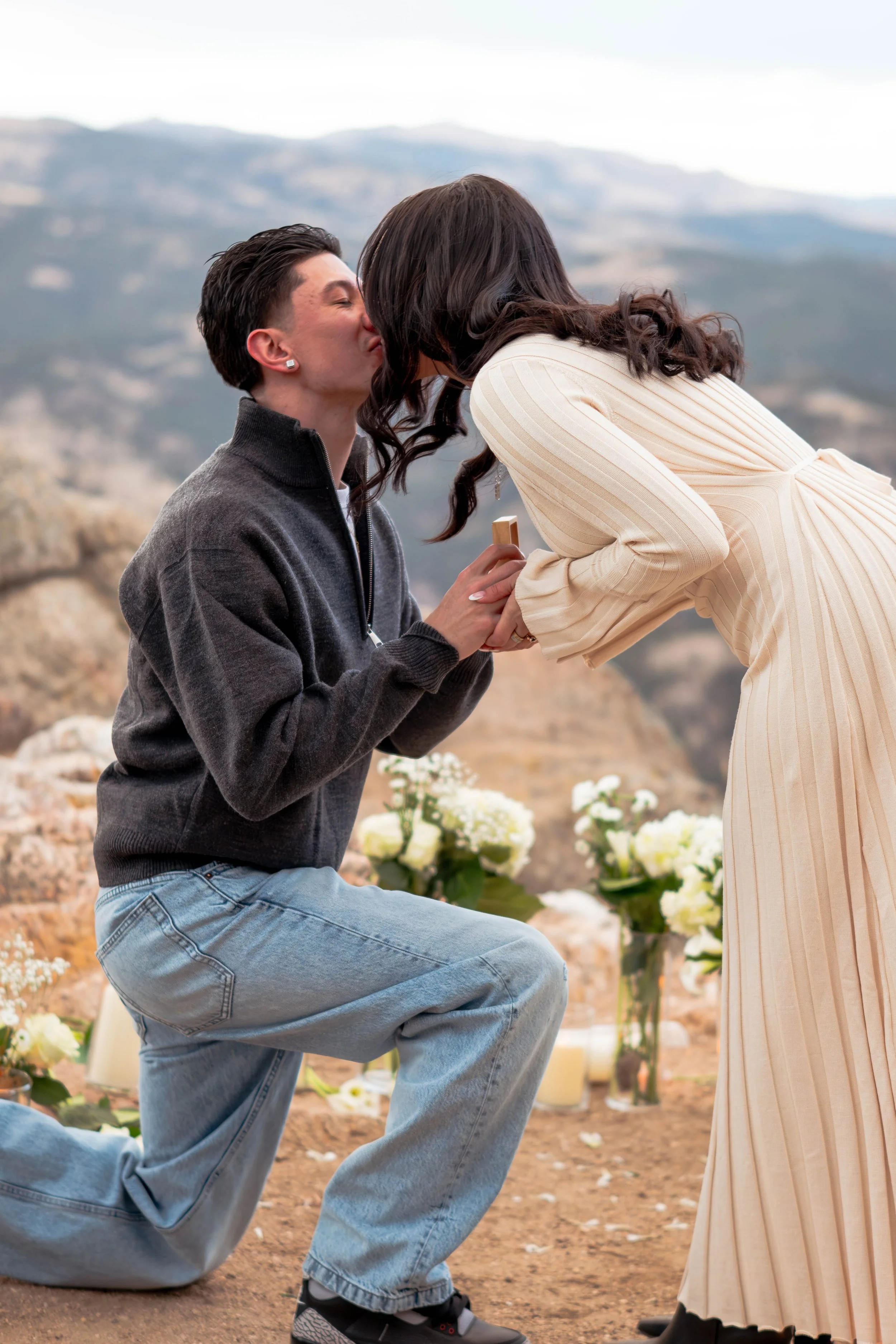 A man kneels on one knee proposing marriage to a woman who is leaning forward, showing affection, outdoors with mountains and flowers in the background.