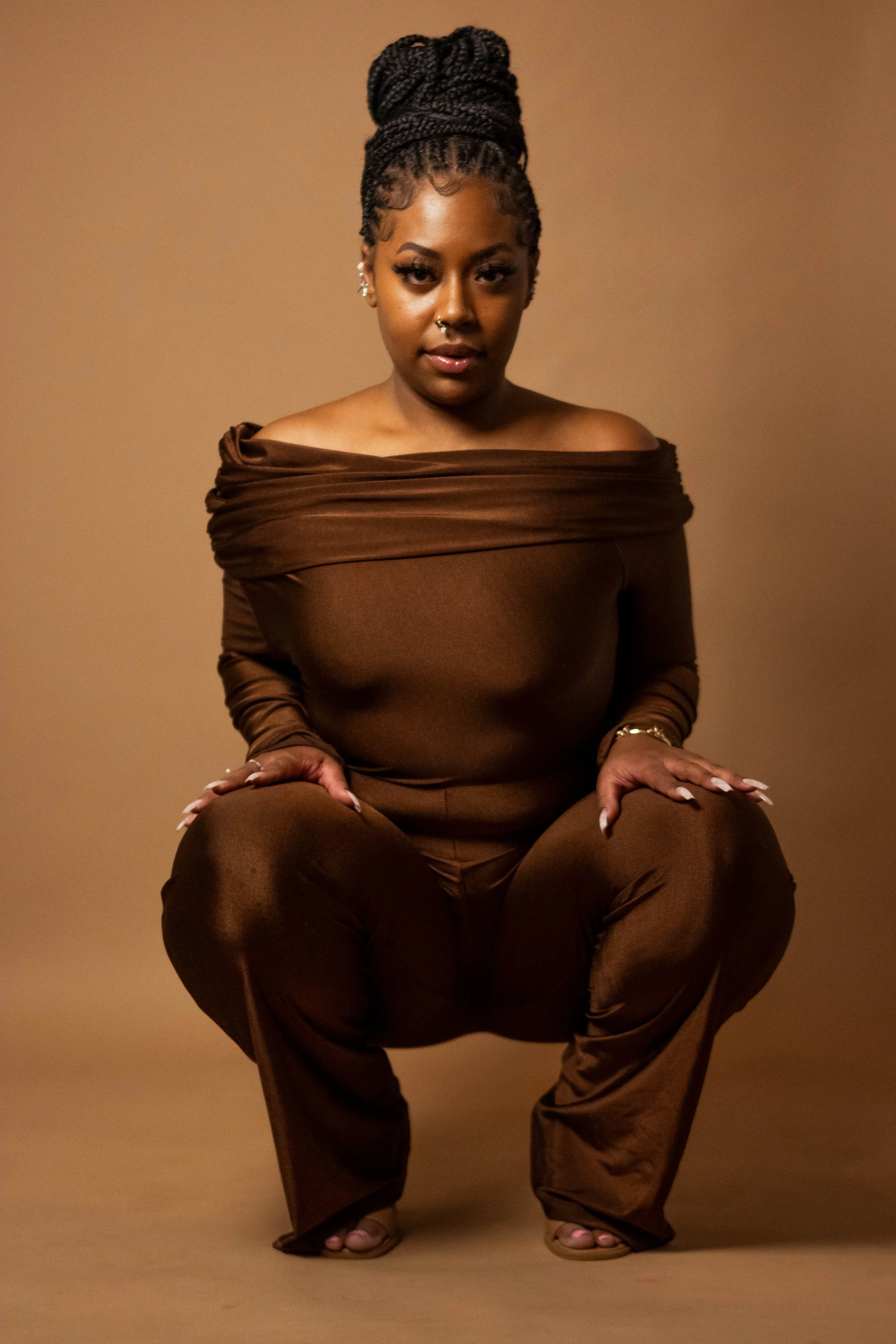 A woman with braided hair styled in a high bun, wearing an off-the-shoulder brown top and matching pants, squatting in front of a beige background.