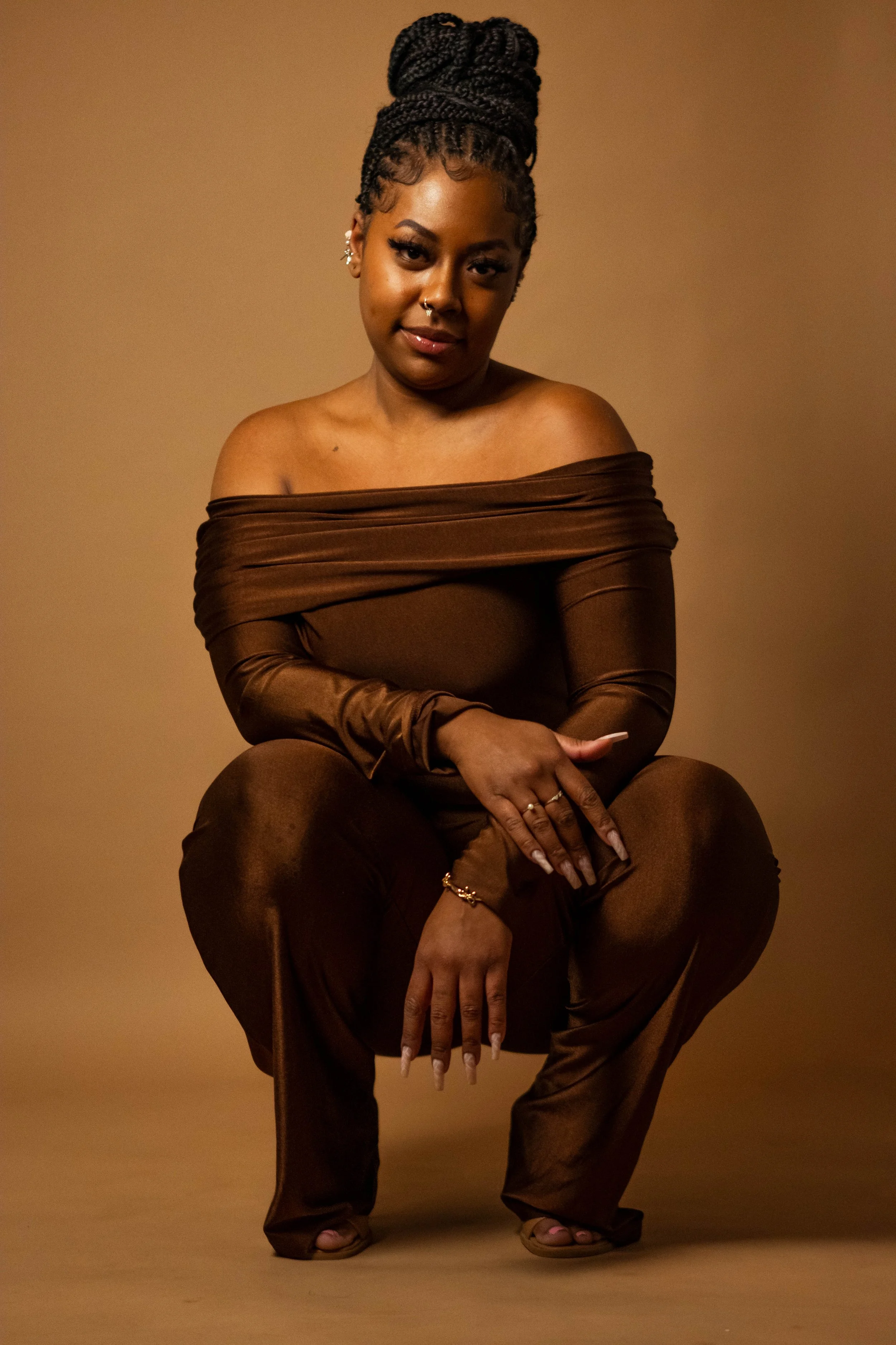 A woman with braided hair styled in a high bun, wearing a brown off-the-shoulder outfit, squatting against a brown background with a confident expression.