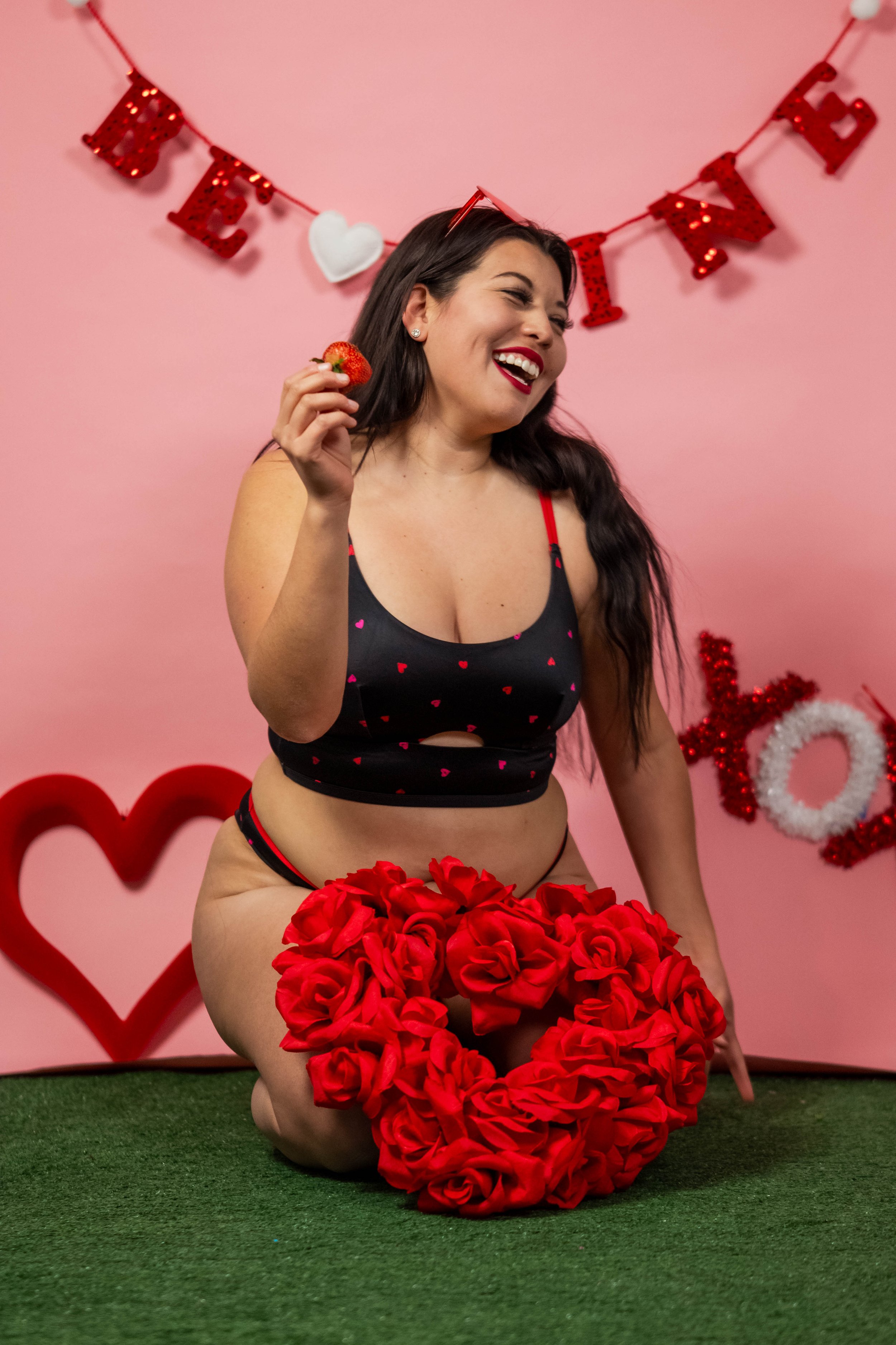 A woman with long dark hair, wearing a black crop top with pink hearts and red straps, is kneeling on green grass in front of a pink background. She is holding a strawberry, smiling, and has a large heart-shaped decoration made of red roses in front 