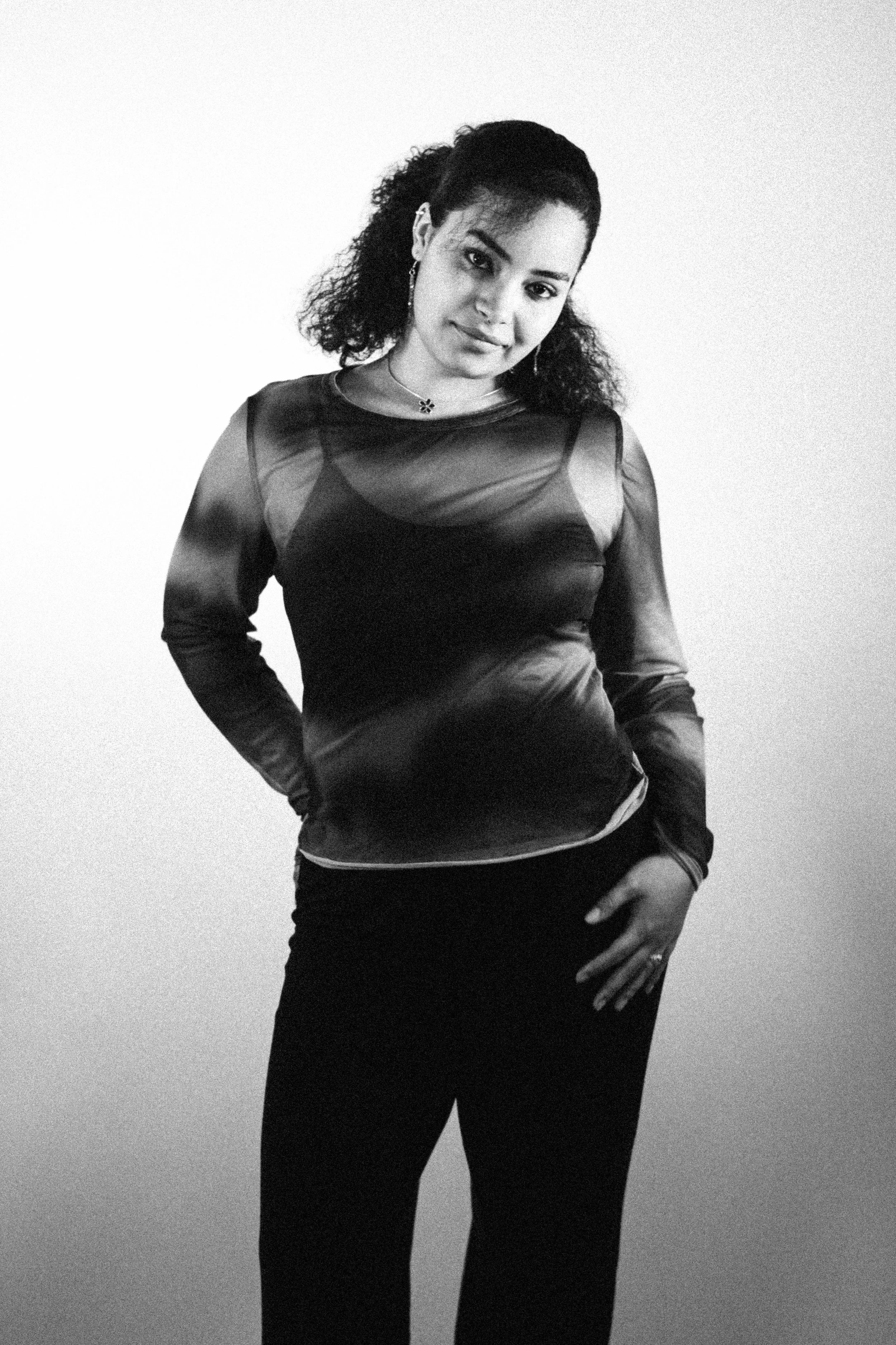 Black and white photo of a woman with curly hair wearing a sheer long-sleeve top and dark pants, posing with hands on hips against a plain background.