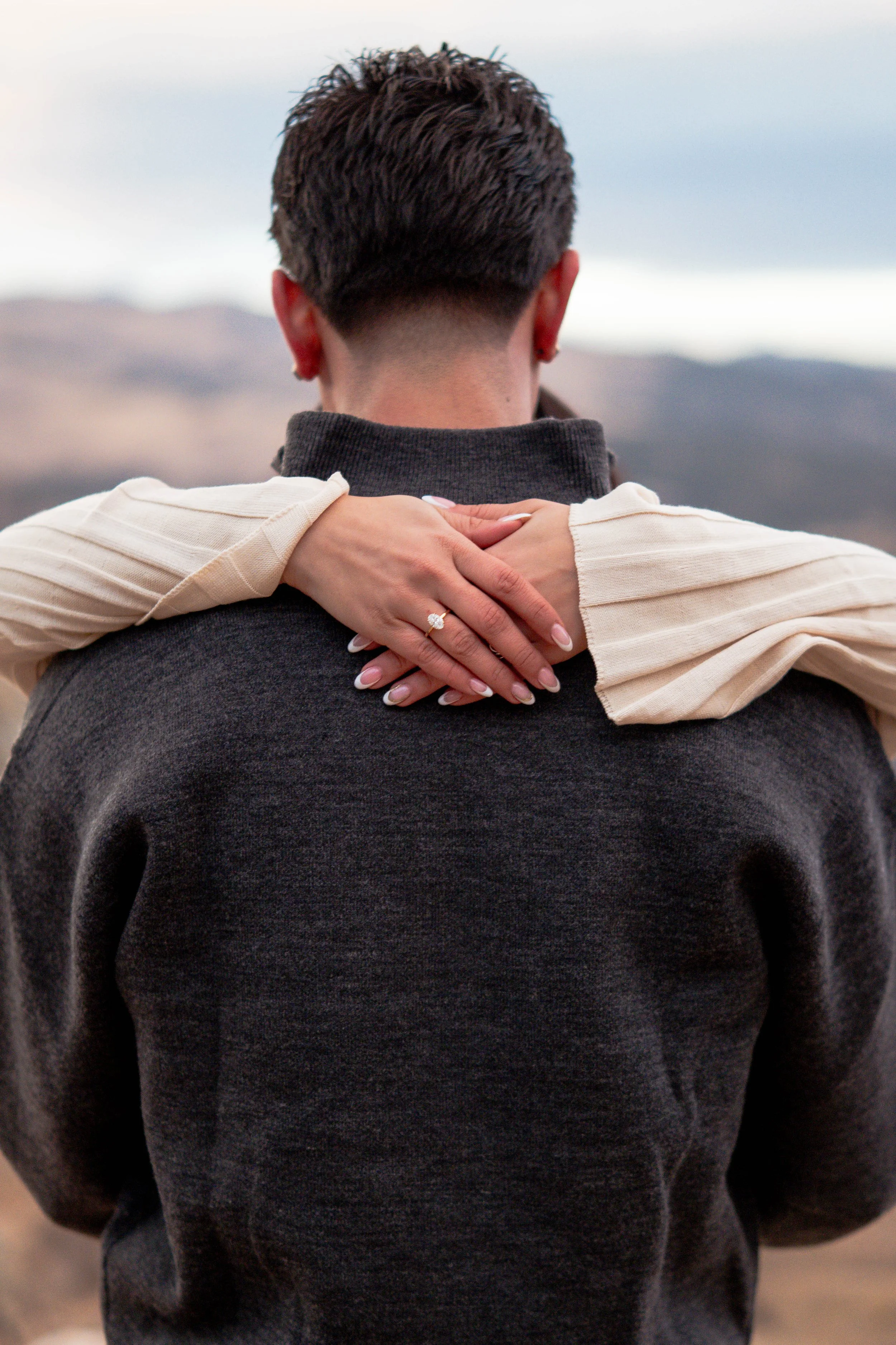 A person with short dark hair, seen from the back, wearing a dark sweatshirt, being hugged from behind by someone with long, light-colored sleeves, with an engagement ring on their finger.